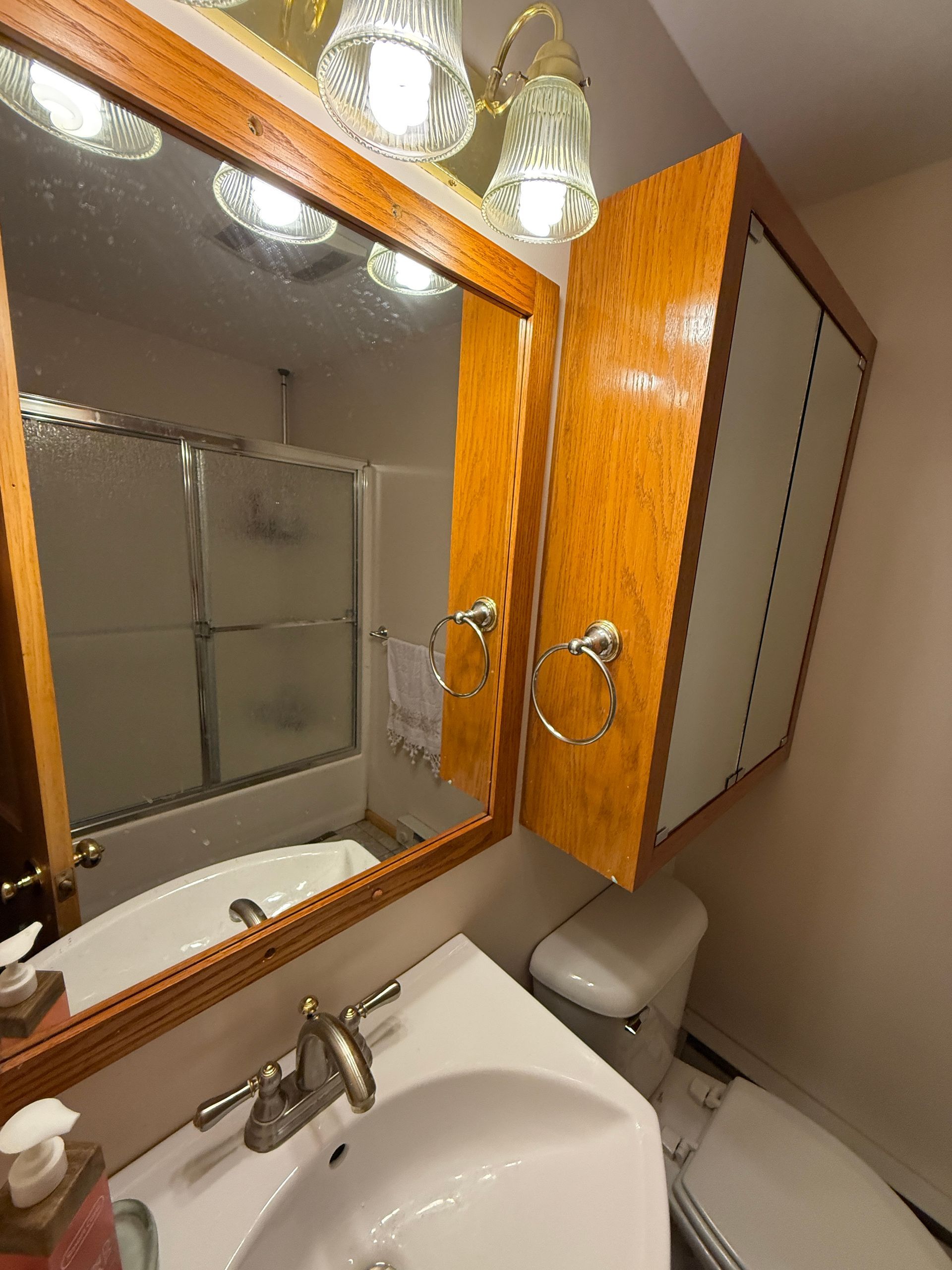Bathroom mirror and cabinet above sink, toilet to the right, shower in the mirror's reflection.