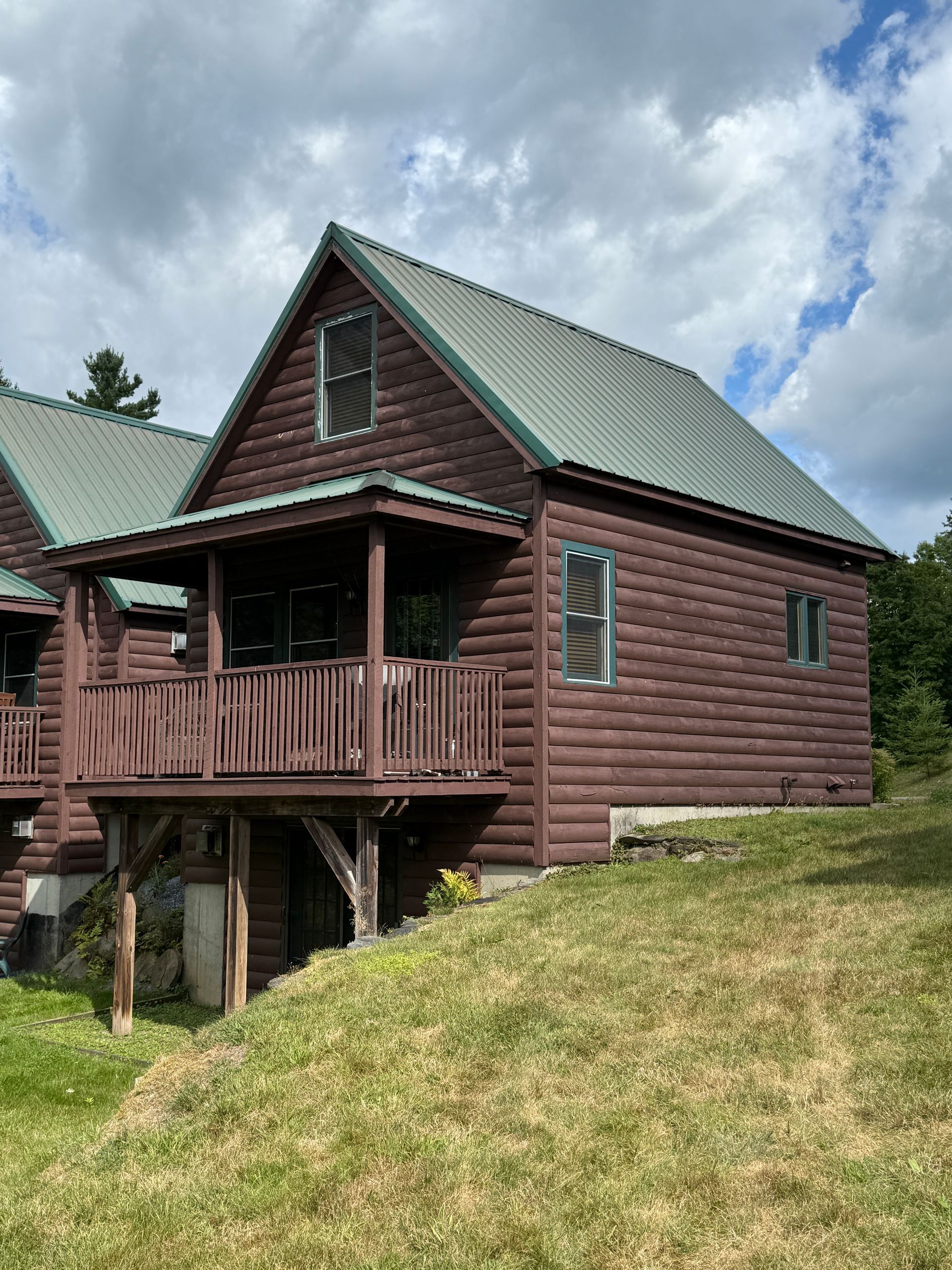 Brown log cabin with green roof and small porch, set on a grassy hill under a cloudy sky.