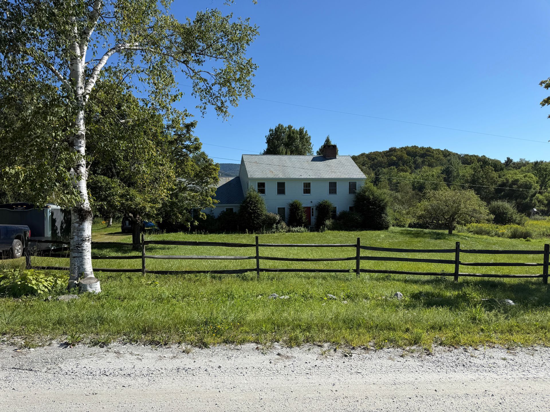 A two-story white house with a gray roof and a wooden fence, set against a green field and a sunny blue sky.