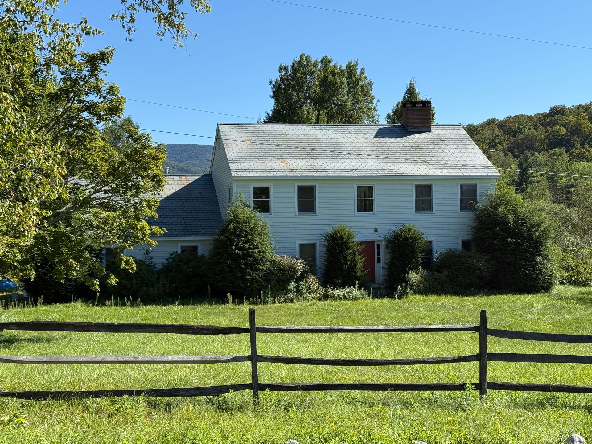 Two-story white house with a gray roof and red door, behind a wooden fence, under a blue sky.