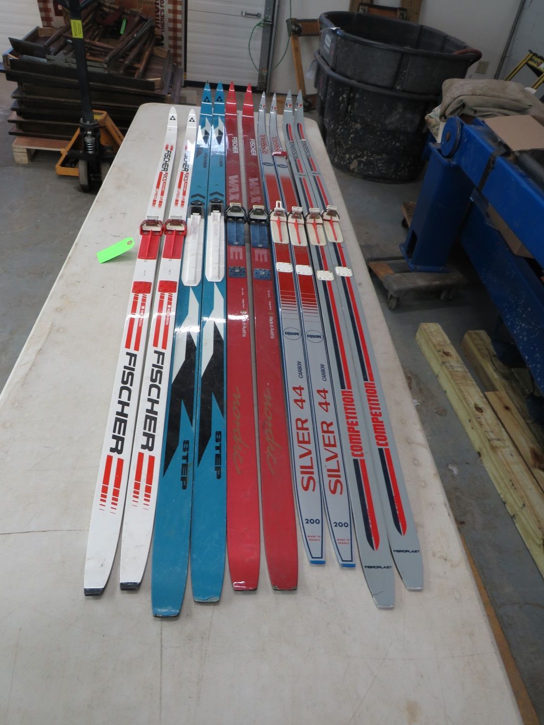 Assortment of cross-country skis, mostly red, blue, and white, on a table in a workshop setting.