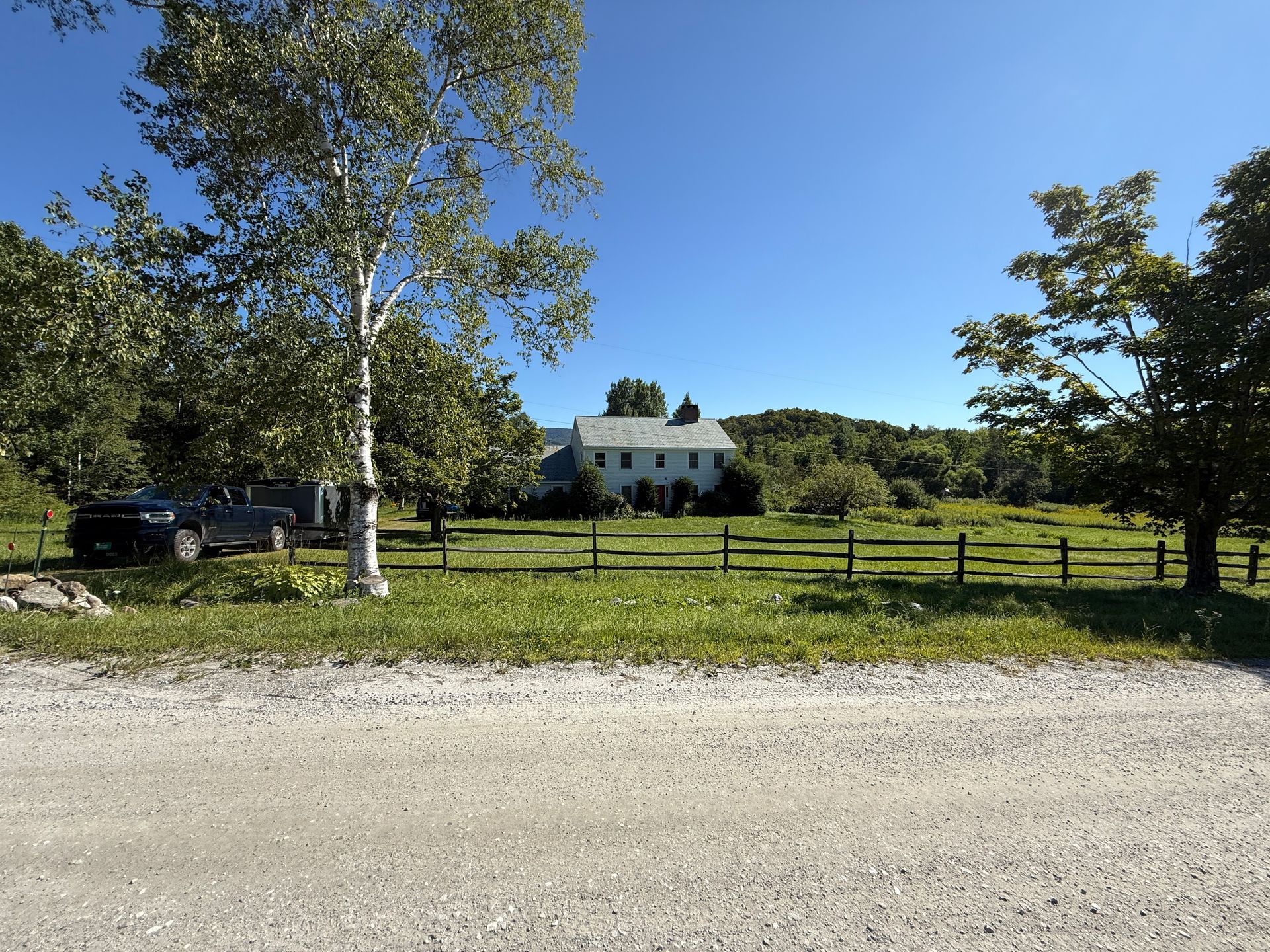 House behind a grassy field with a wooden fence. Trees frame the scene under a clear blue sky.