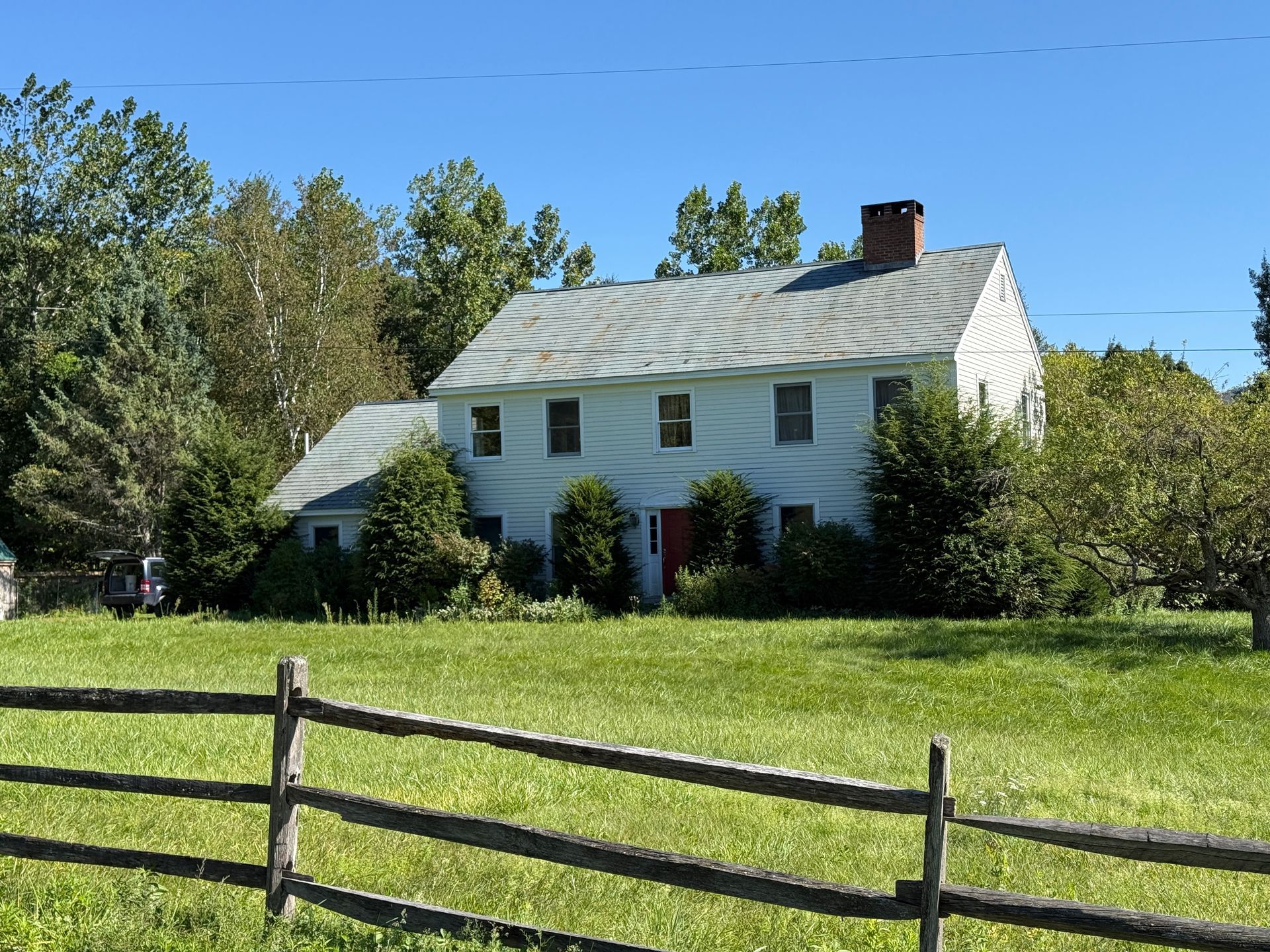 White two-story house with gray roof, red door, and chimney, behind a wooden fence in a grassy yard under a blue sky.