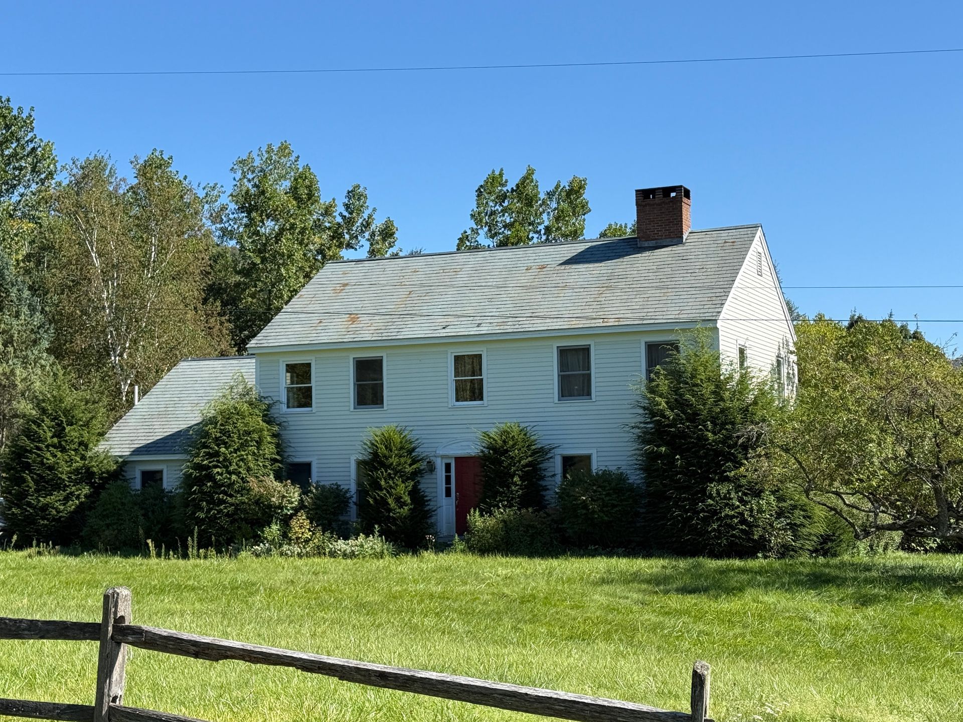 White two-story farmhouse with a metal roof, red door, and chimney, set in a field.