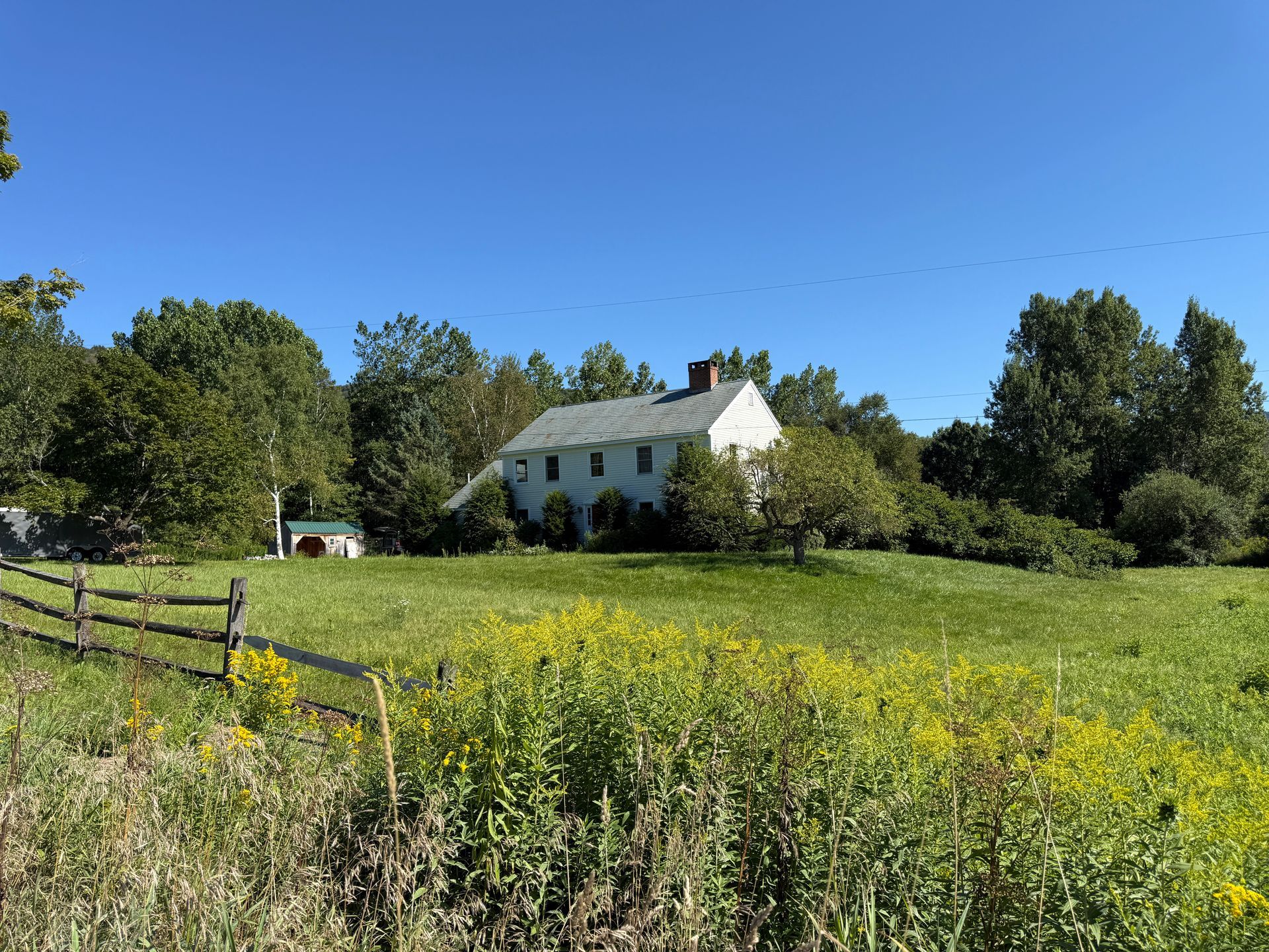White house with a chimney in a green field, surrounded by trees and a wooden fence, under a blue sky.
