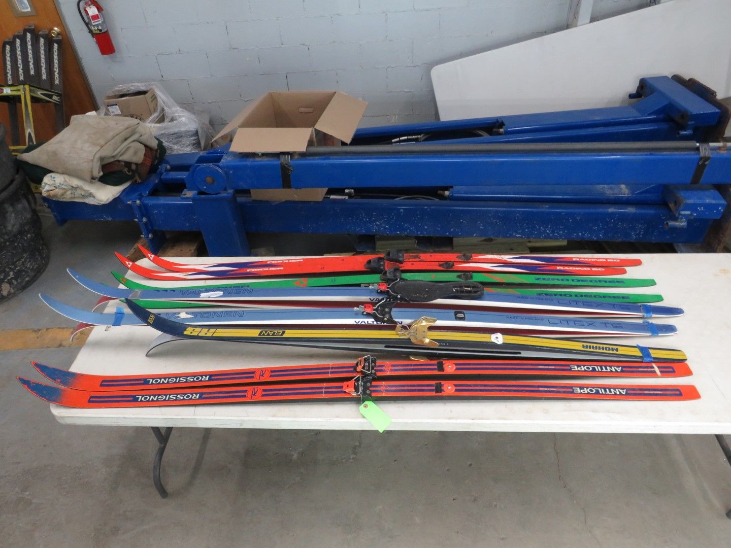 A stack of cross-country skis, various colors, on a white table. Blue machine in the background.