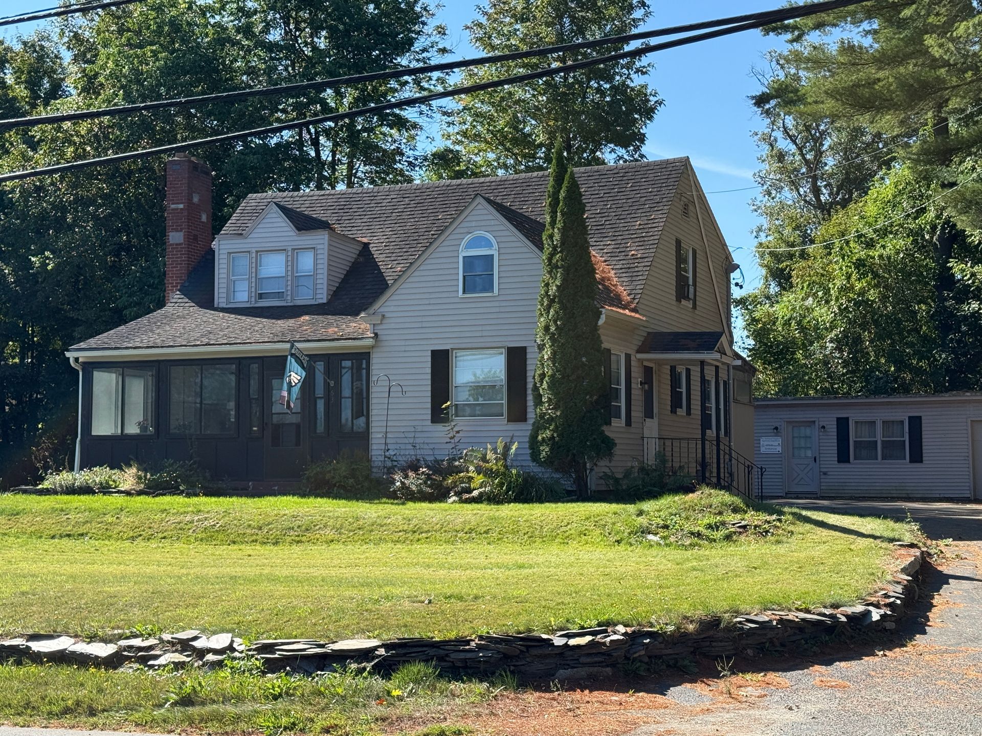 Cottage-style two-story house with green lawn and trees, blue sky. Screened porch, chimney.