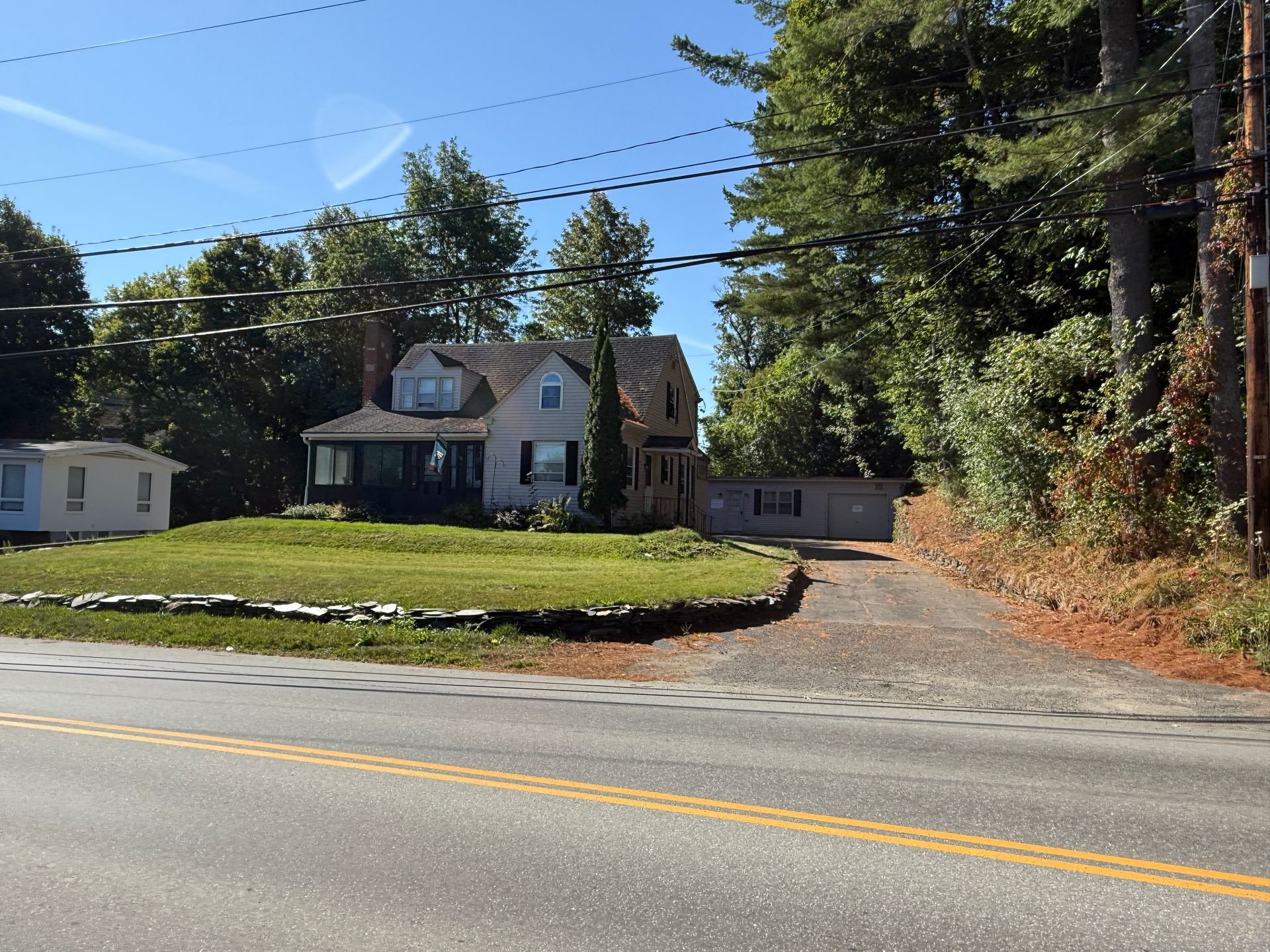 House with light gray siding, dormer, and attached garage sits on a lawn, next to a road, surrounded by trees.