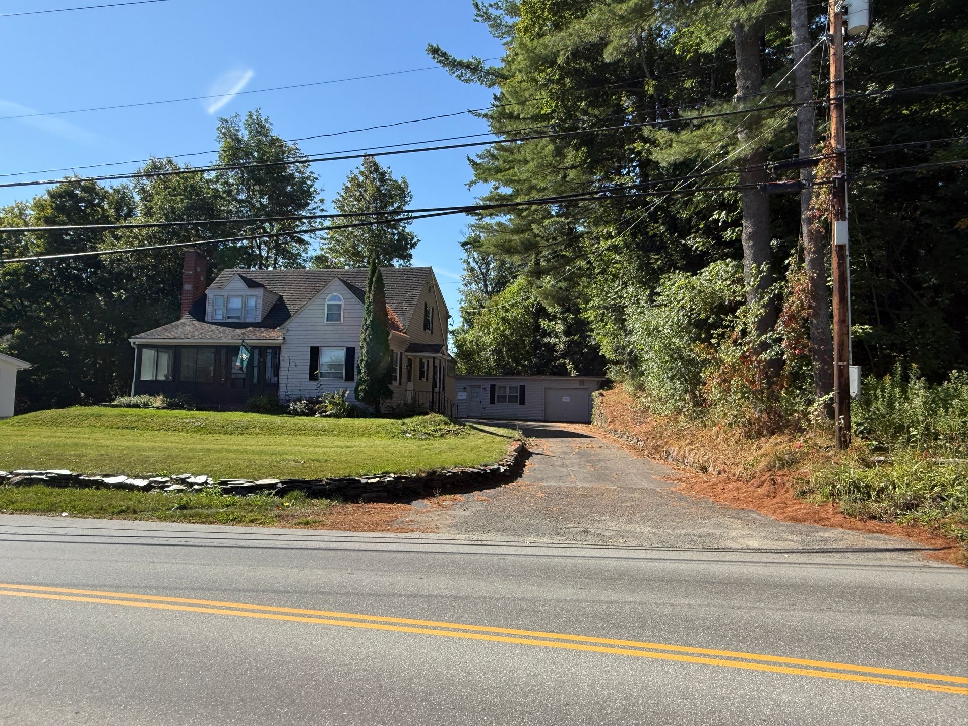 A small two-story house with a driveway off a paved road, surrounded by grass and trees.