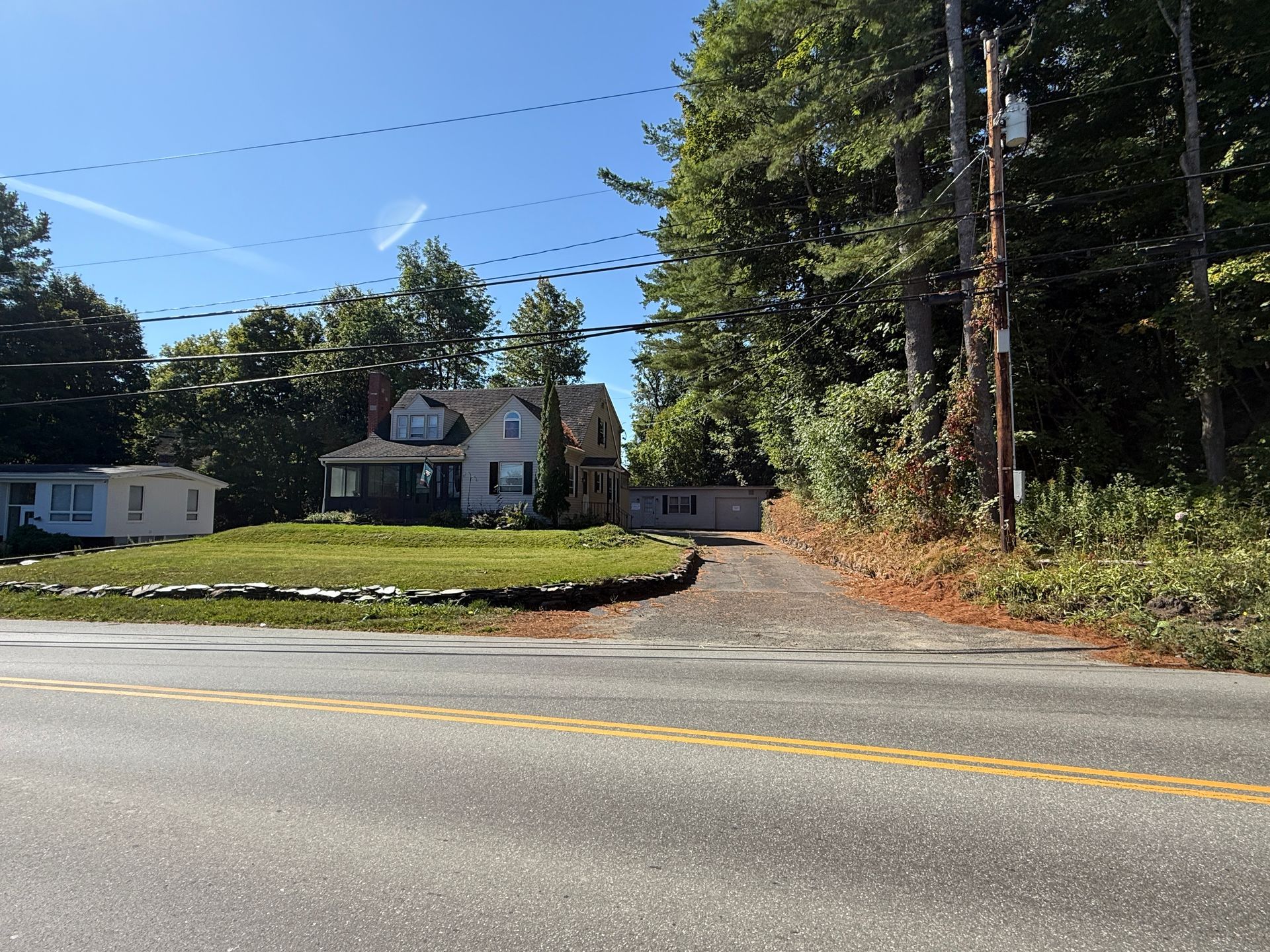 A house with a driveway next to a road, under a blue sky, power lines overhead.