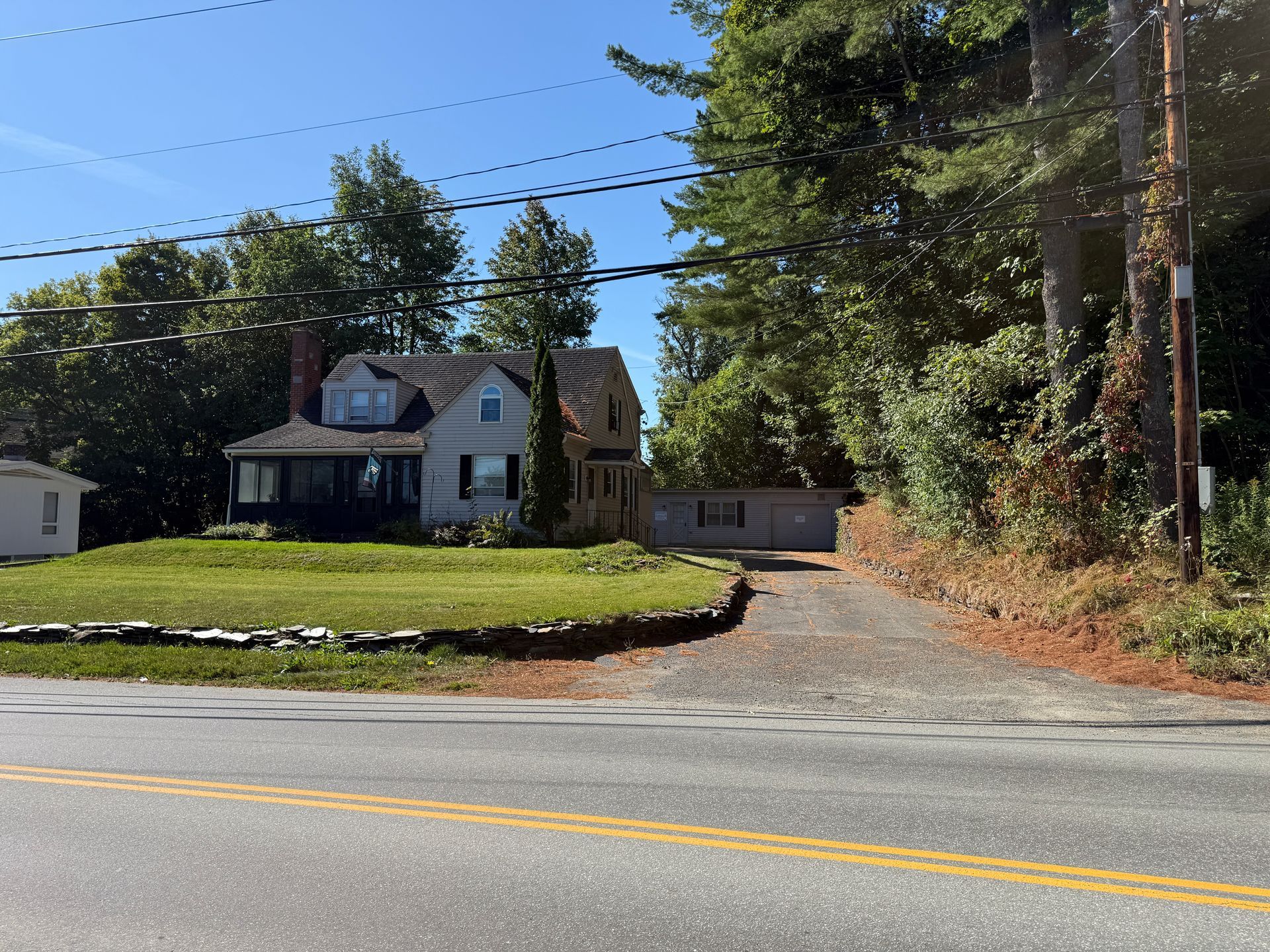 Cottage with gray siding, dormer window, and driveway off a two-lane road, surrounded by trees.