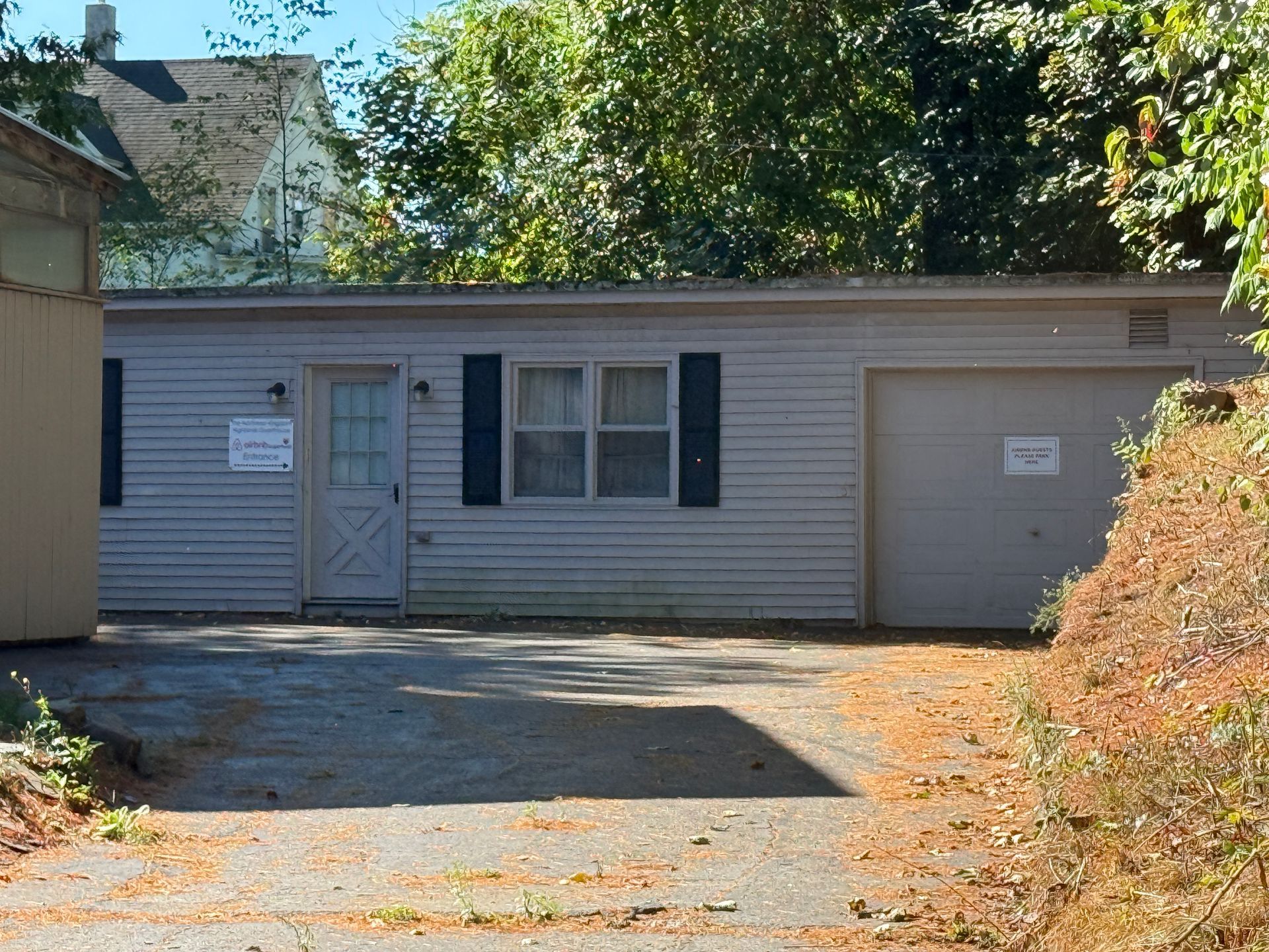 A light blue, one-story building with a garage door and small front door, parked in front of a tree and driveway.