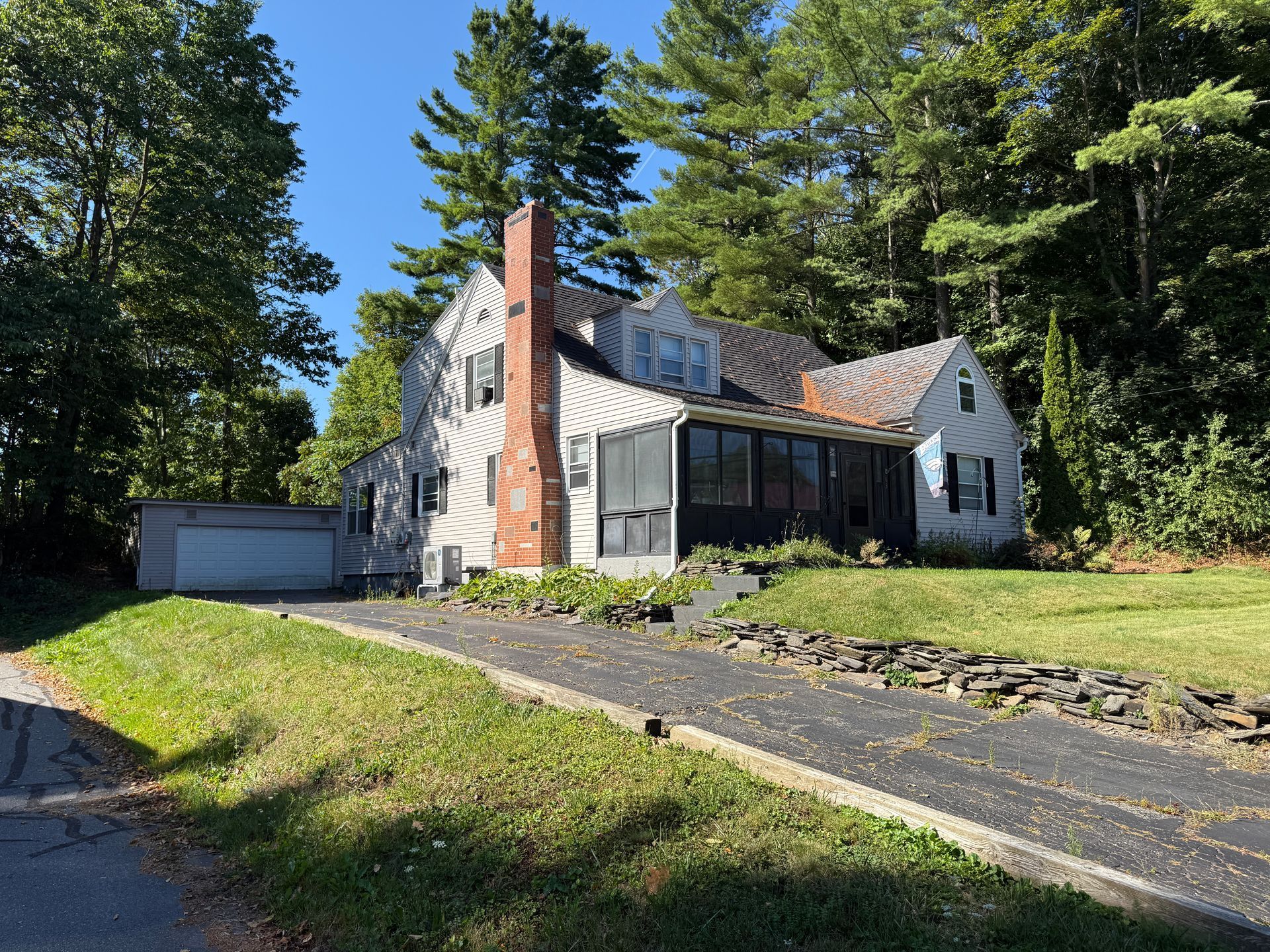 House with a driveway, brick chimney, and attached garage surrounded by green grass and trees on a sunny day.