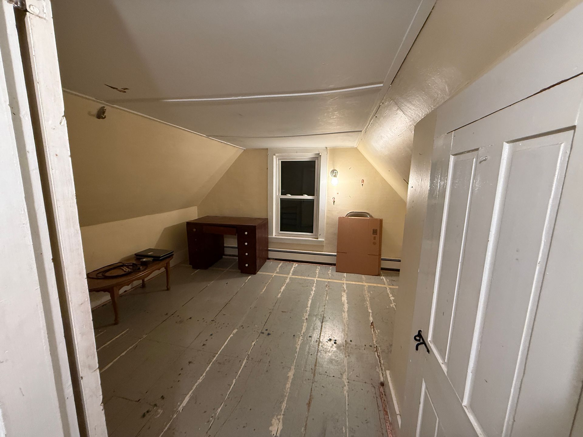 A small attic room with sloped ceilings, a desk, a bench, and a closed door. The walls are yellow.