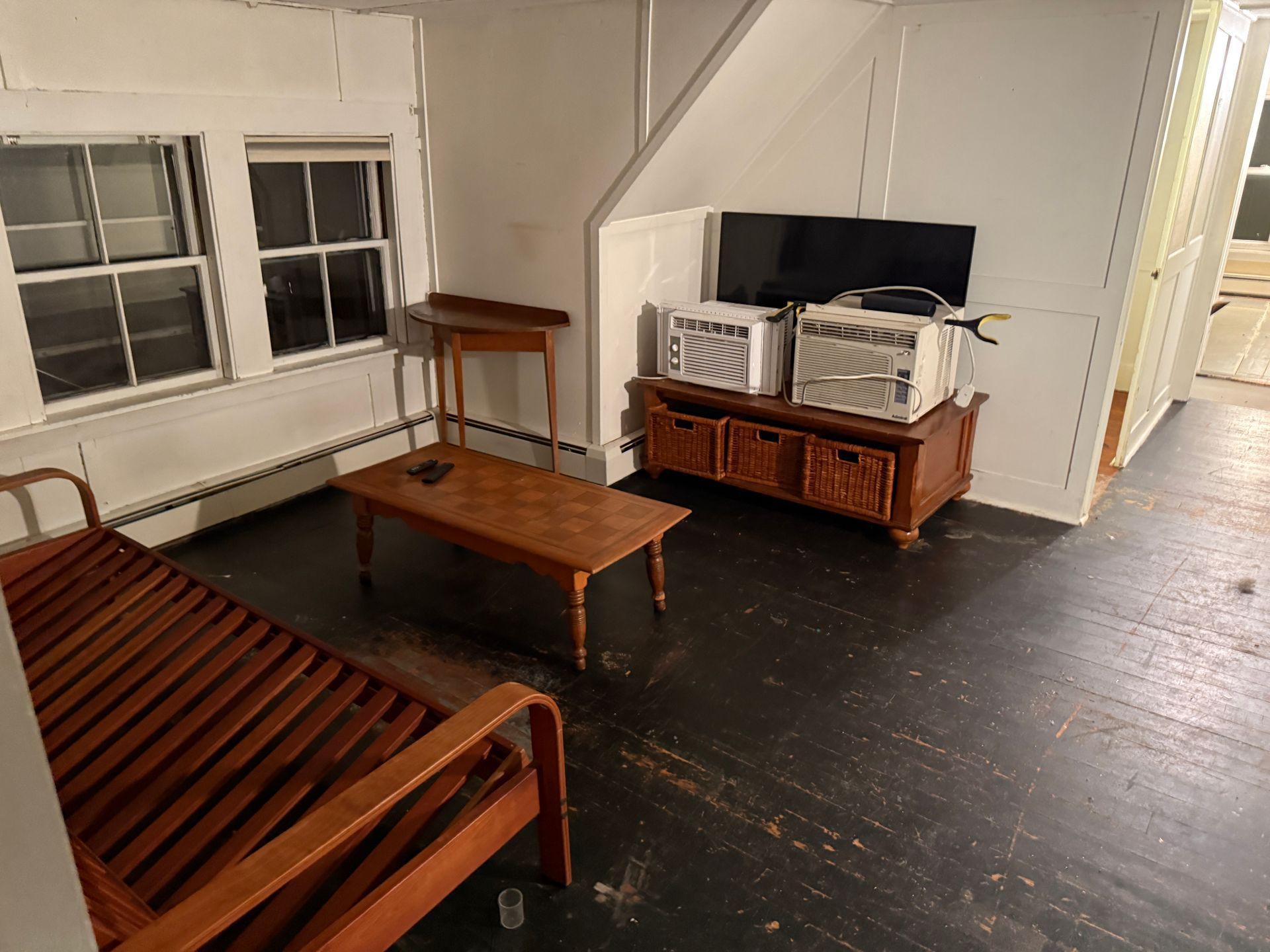 Living room with a futon, TV, coffee table, and windows. Dark wood floors and white walls.
