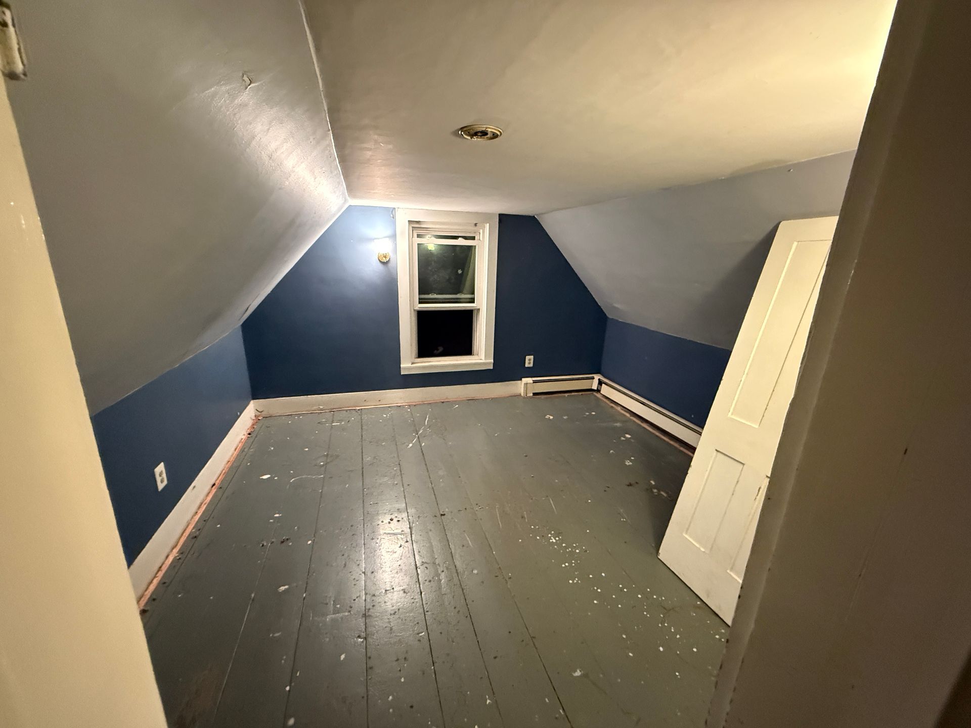 Empty attic room with blue and gray walls, window, and gray wooden floor.