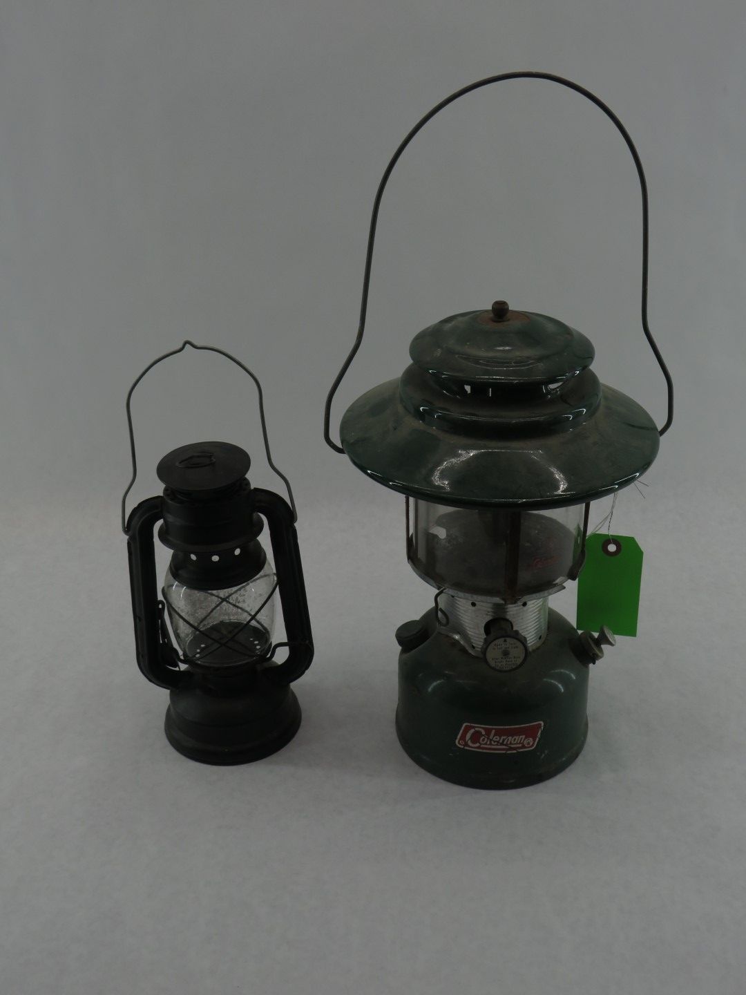 Two camping lanterns: small black one on the left, larger green Coleman on the right, both with wire handles.
