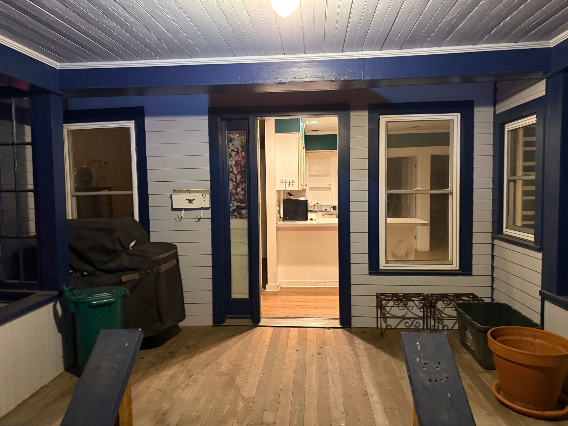 Enclosed porch with blue trim and pale gray siding, open doorway to a kitchen, two windows.