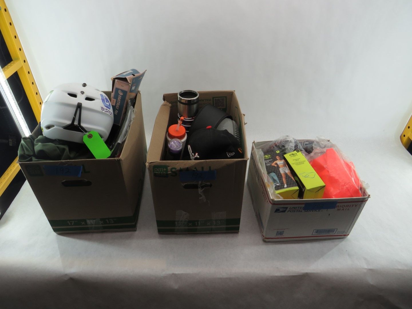 Three cardboard boxes filled with various items, including a helmet, thermos, and packaged goods, on a white surface.