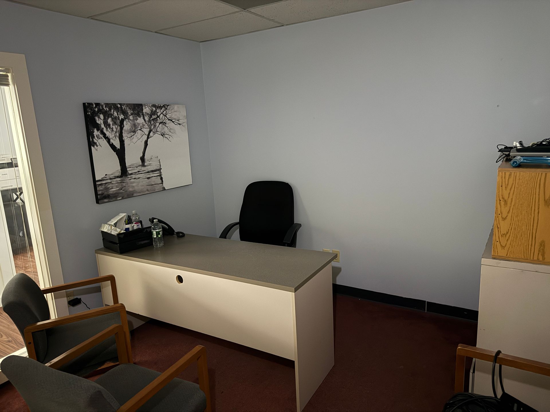 Office with desk, chair, artwork, and two guest chairs. Blue walls and red-brown flooring.
