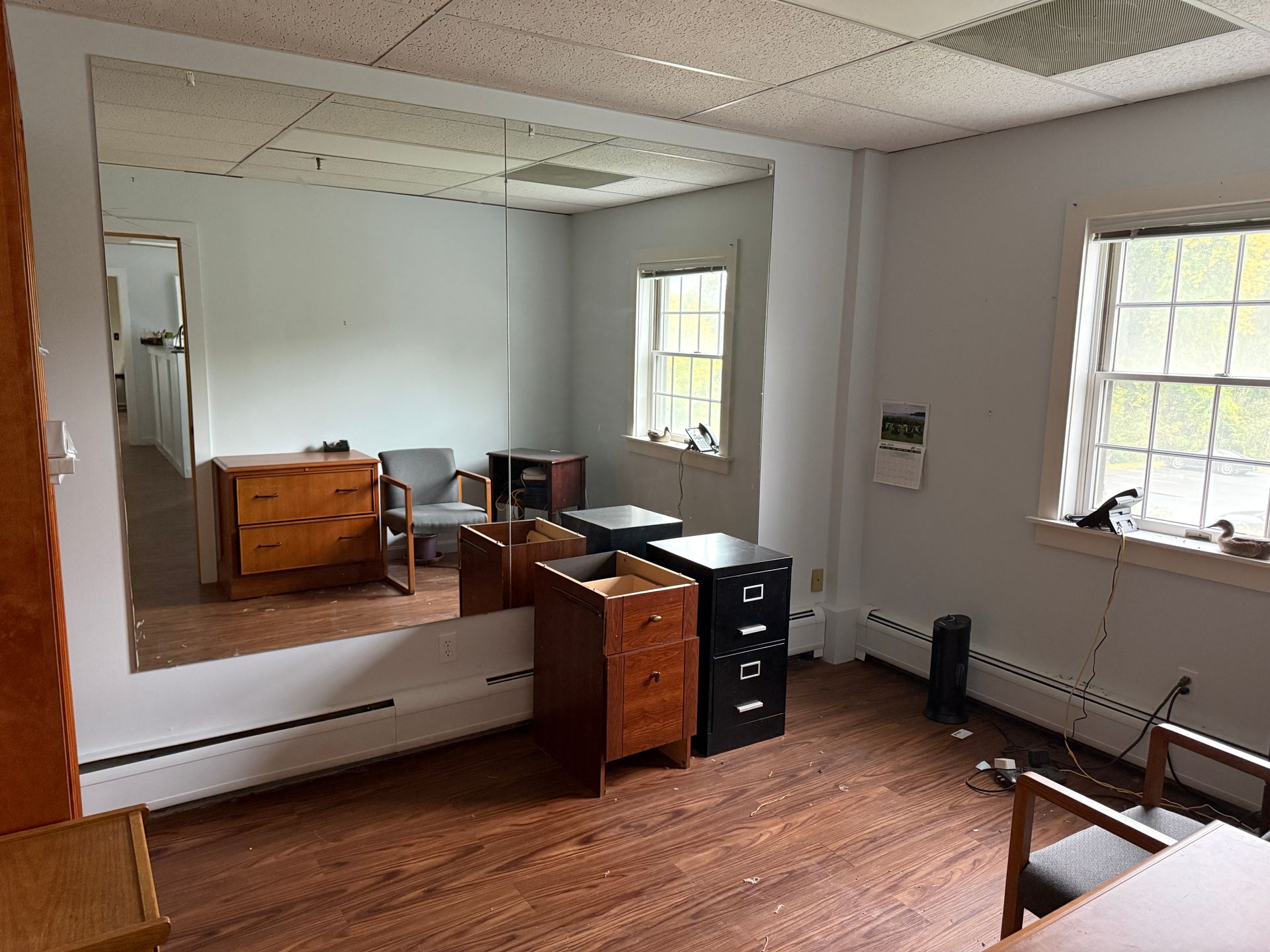 An office room with a large mirror, windows, wooden floors, and various pieces of furniture.