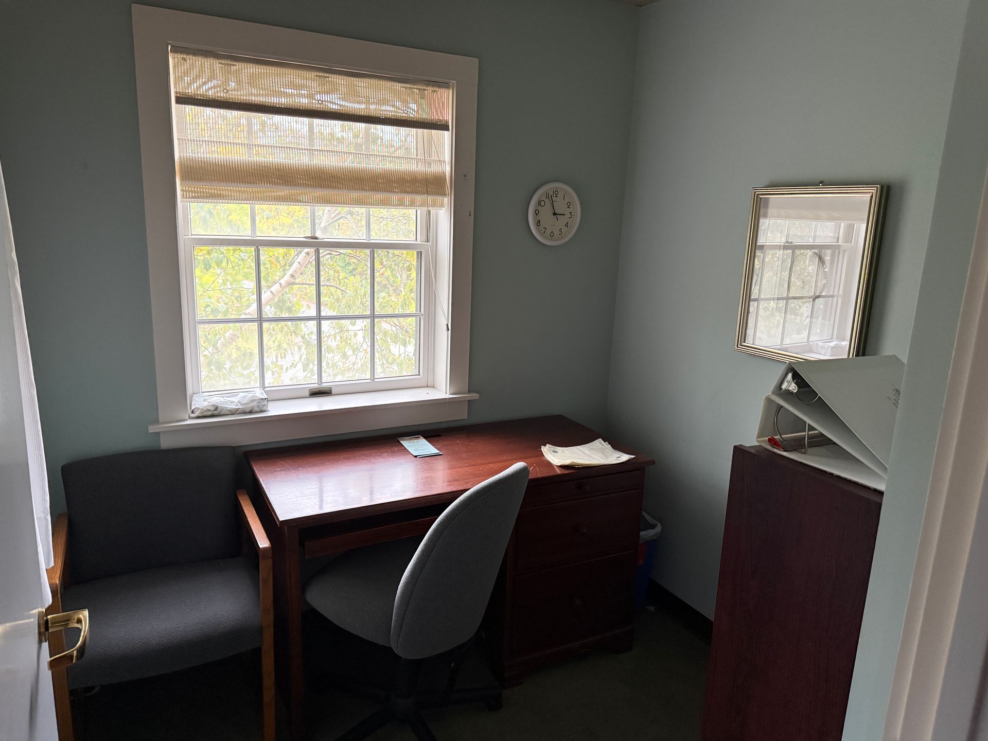 Small office with desk, chair, window, clock, and mirror; teal walls, brown desk and blinds.