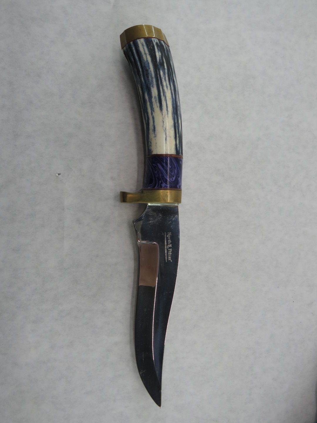 Knife with blue, white, and brass handle. The blade is black with a metallic clip attached.
