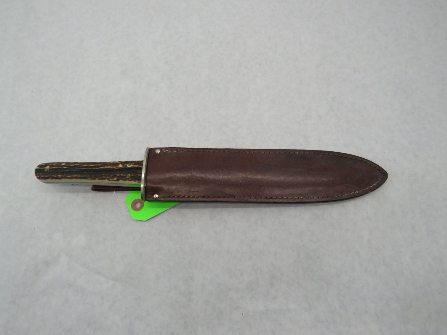 Hunting knife in leather sheath, handle showing, on a white surface. Brown sheath and handle.