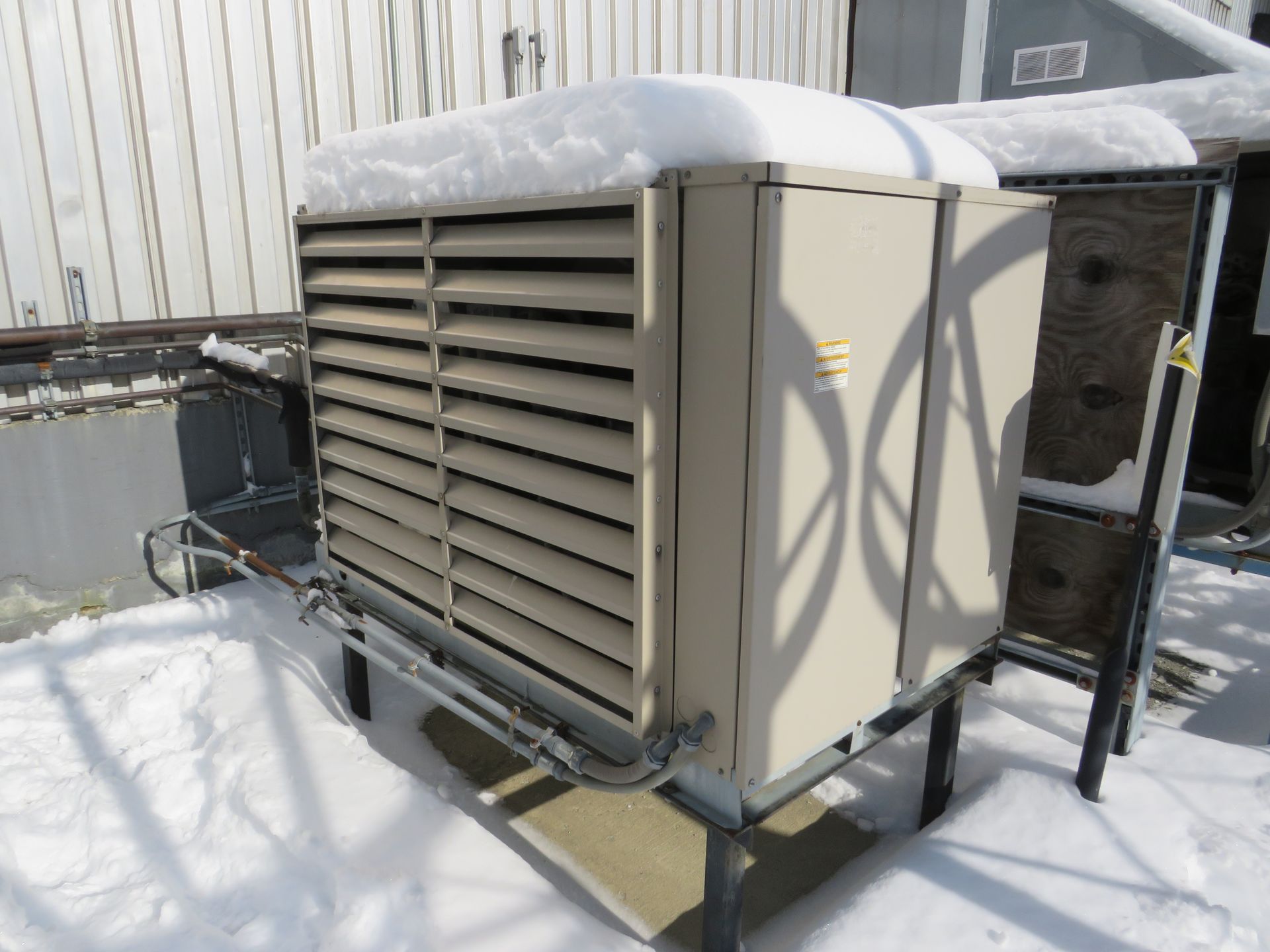 Outdoor air conditioning unit covered in snow, beige metal, on a metal stand, next to a building.