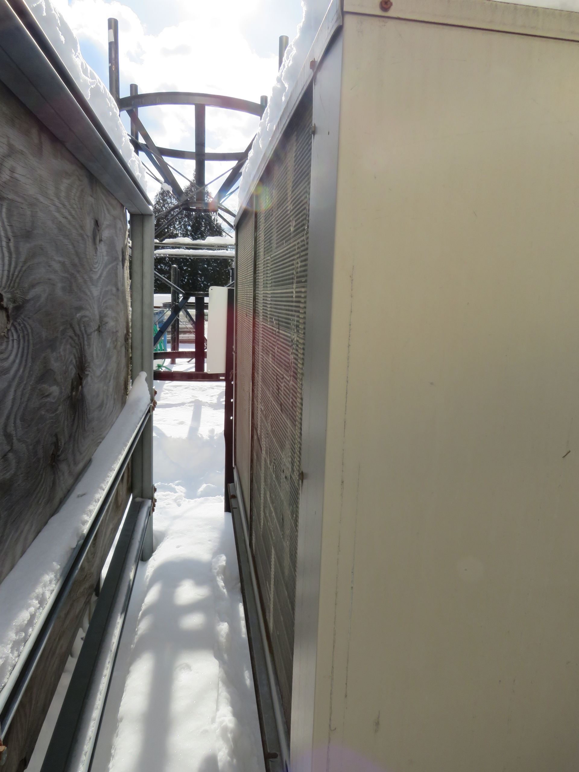 Narrow snow-covered alley between two beige utility structures with a ski lift visible in the distance.