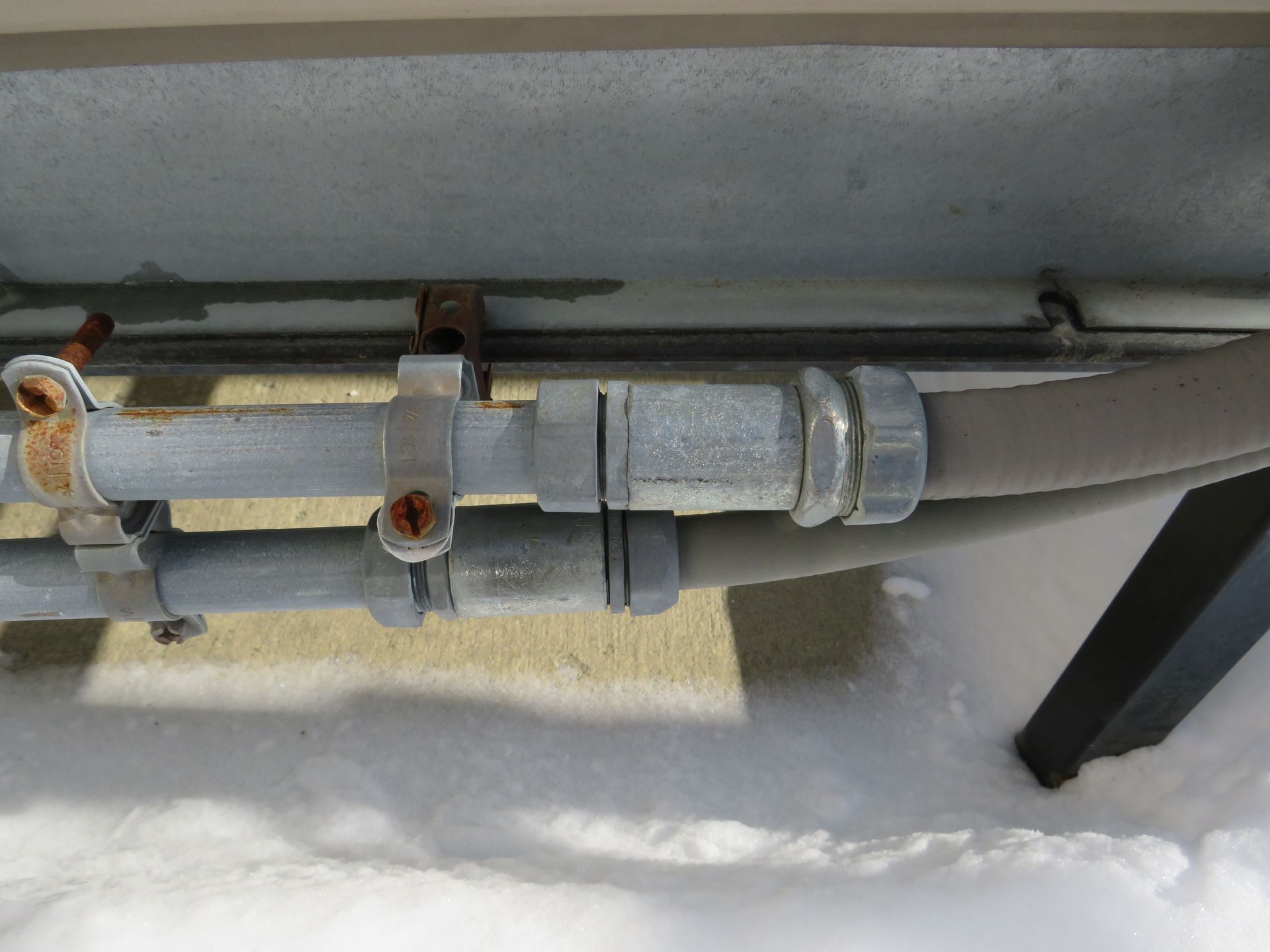 Galvanized metal electrical conduit and cable supported by metal clamps on a snowy exterior wall.