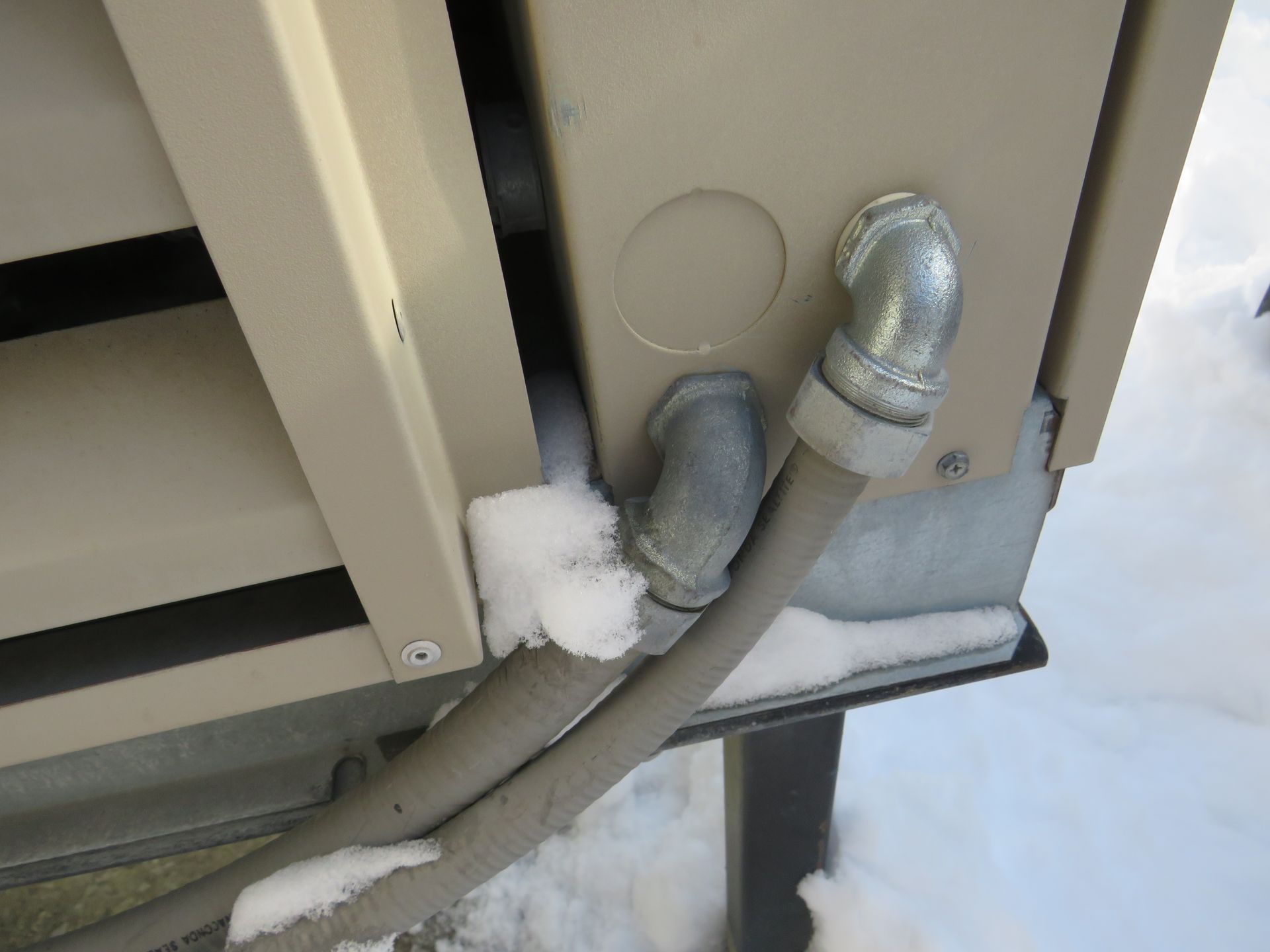 Snow-covered outdoor electrical conduit and junction box. Beige metal, gray conduit, galvanized elbow, snowy setting.