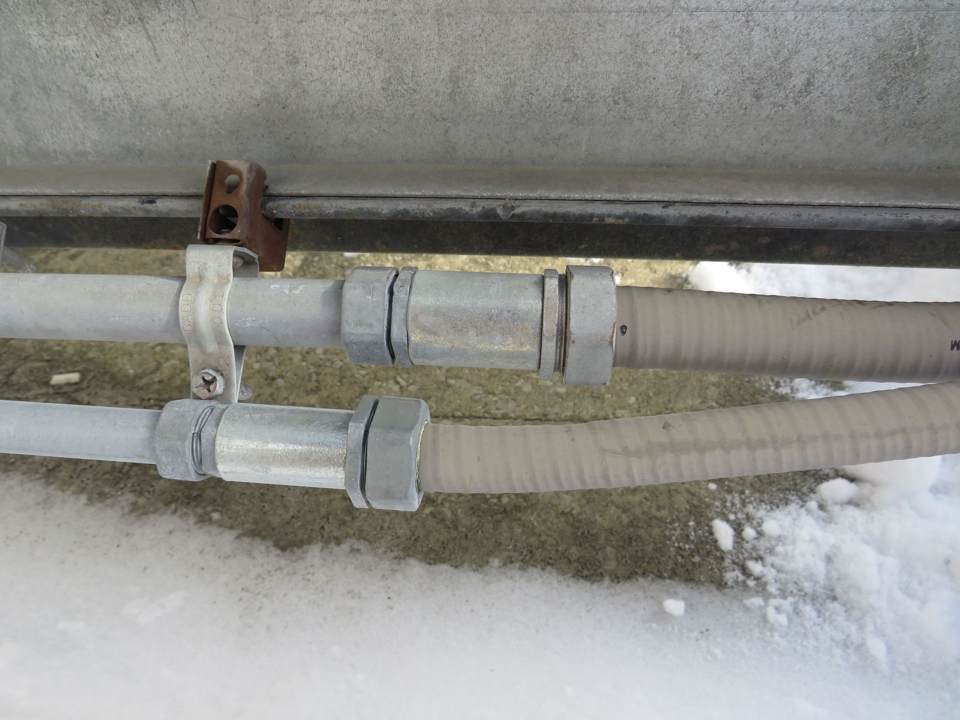 Two conduits, secured with metal clamps, on snow-covered ground, under a metal structure.