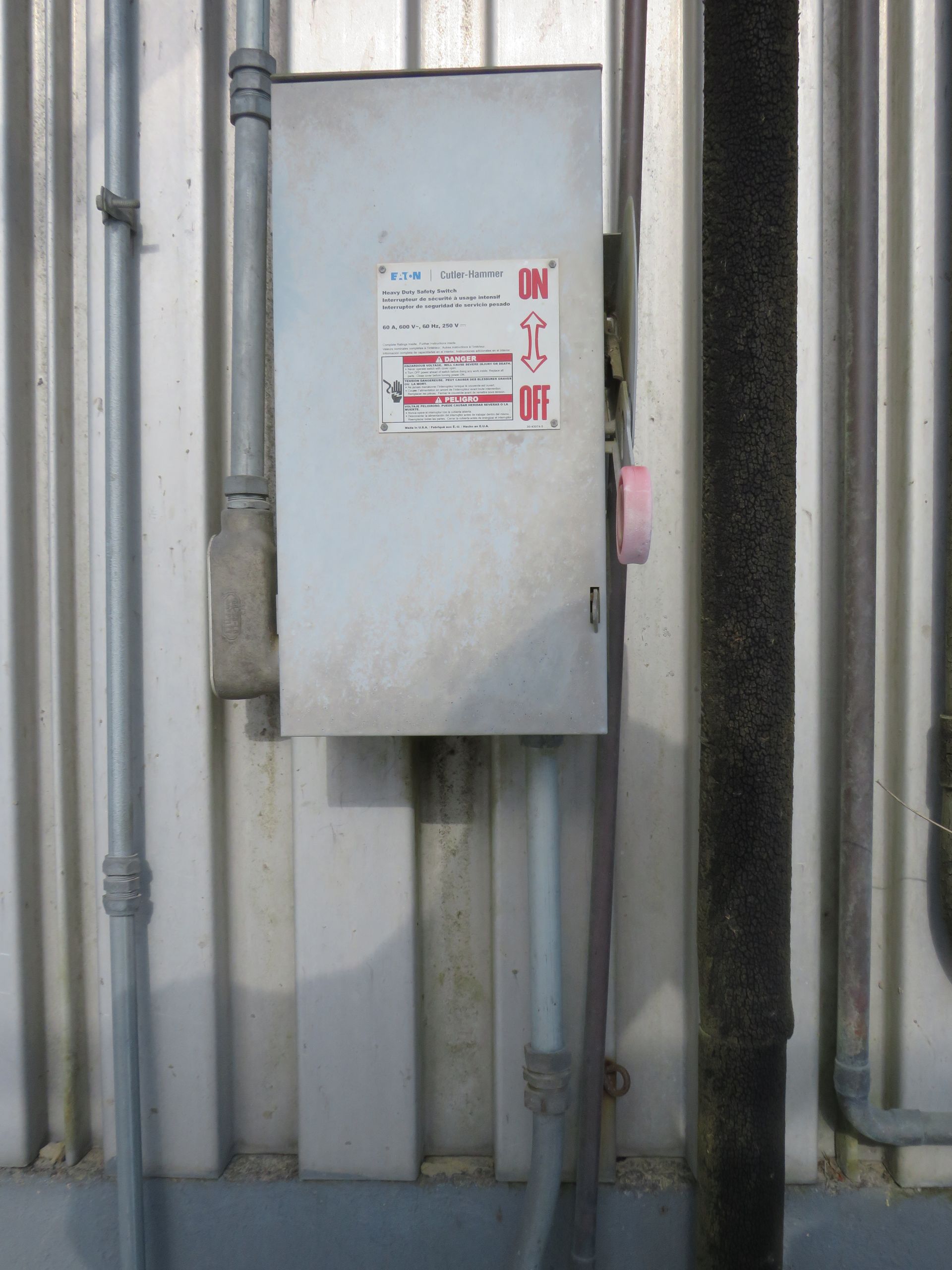 Metal electrical box on a corrugated metal wall, with conduits and a wrapped pipe nearby.