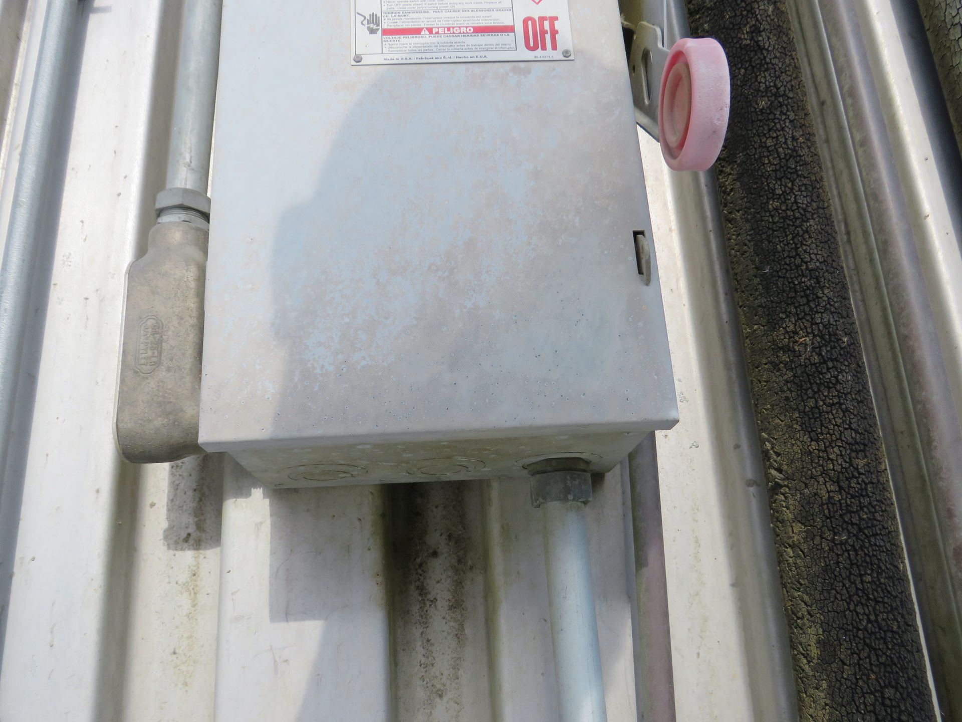 Gray electrical box mounted on a corrugated metal surface with conduit pipes.