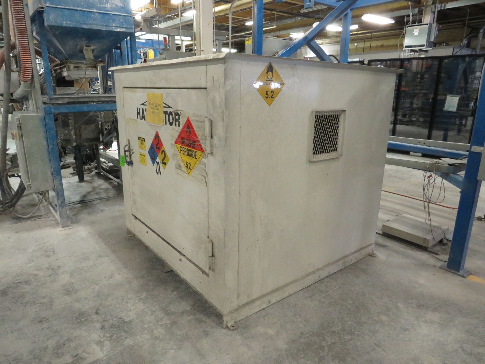 A white hazardous material storage locker in a factory setting, with hazard warning labels.