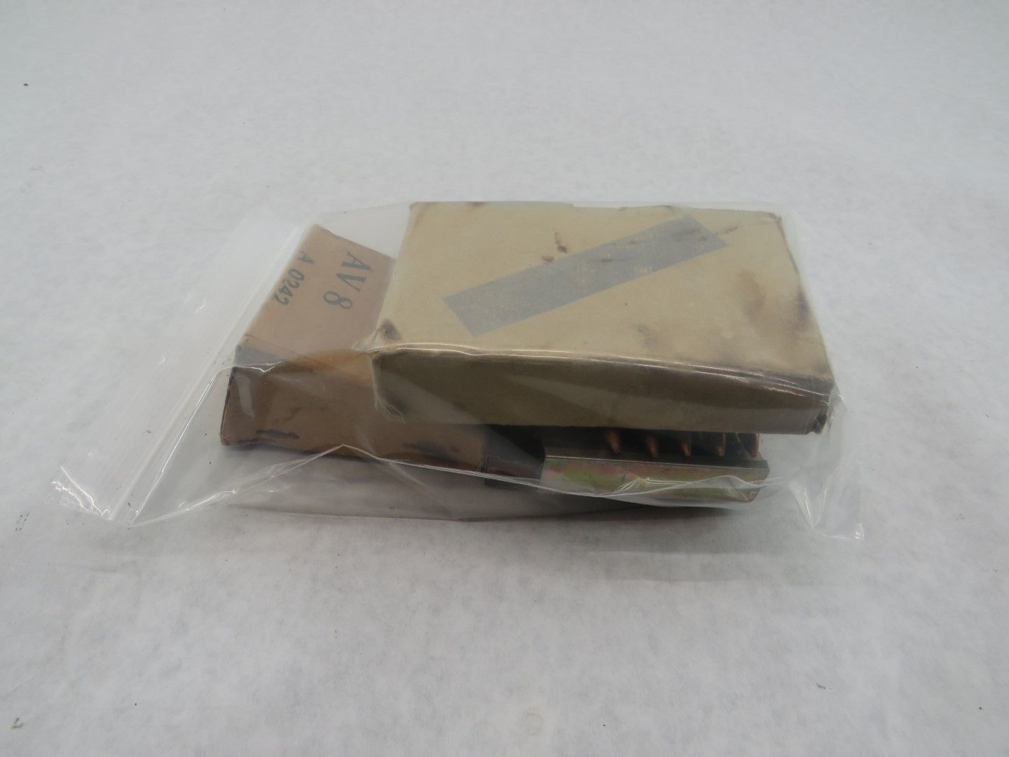 Packaged rectangular object in a clear plastic bag on a white surface. Beige paper and metal components visible.