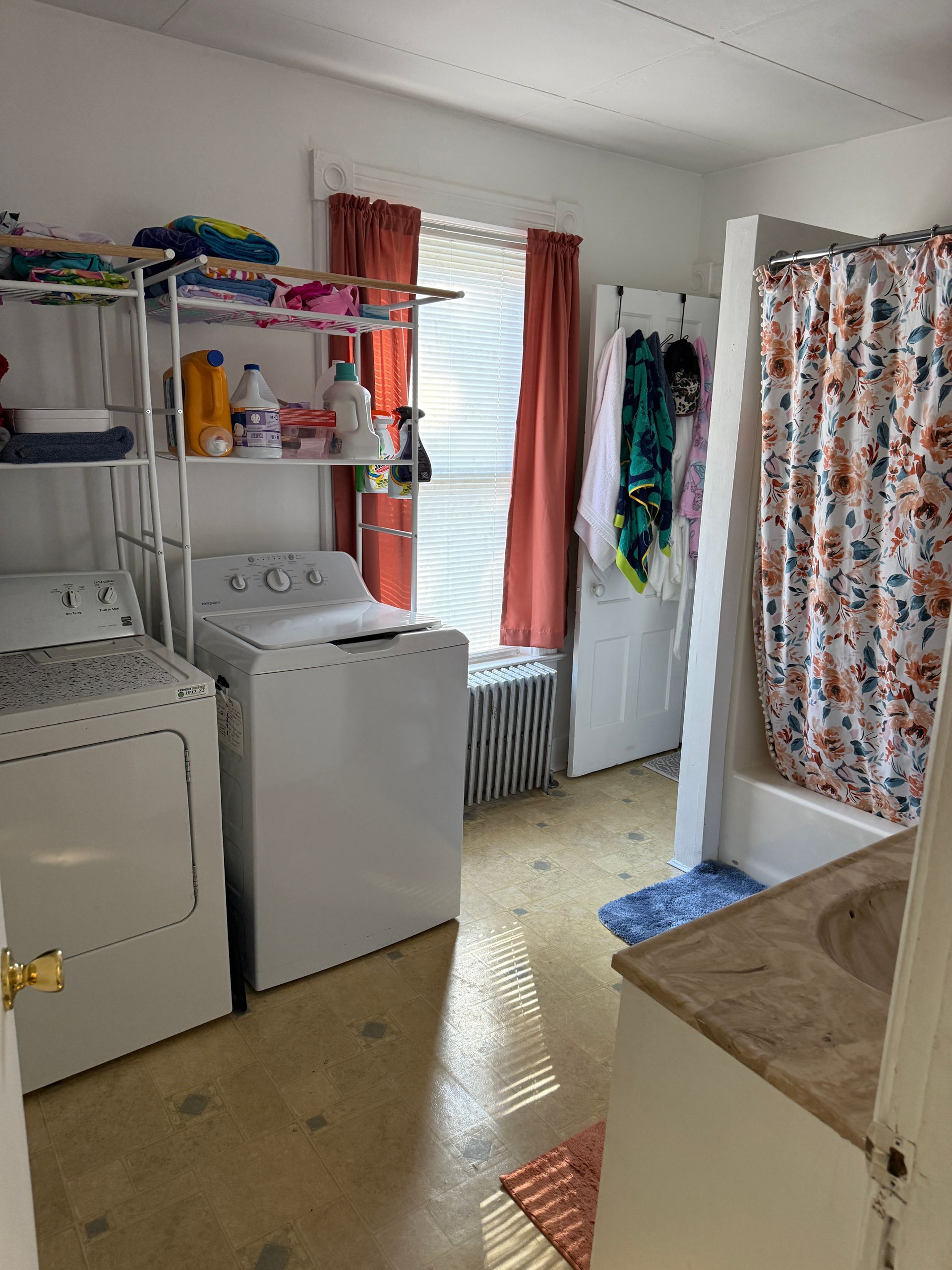 Laundry room with washer, dryer, shelf with supplies, window with blinds, and bathroom with shower curtain.