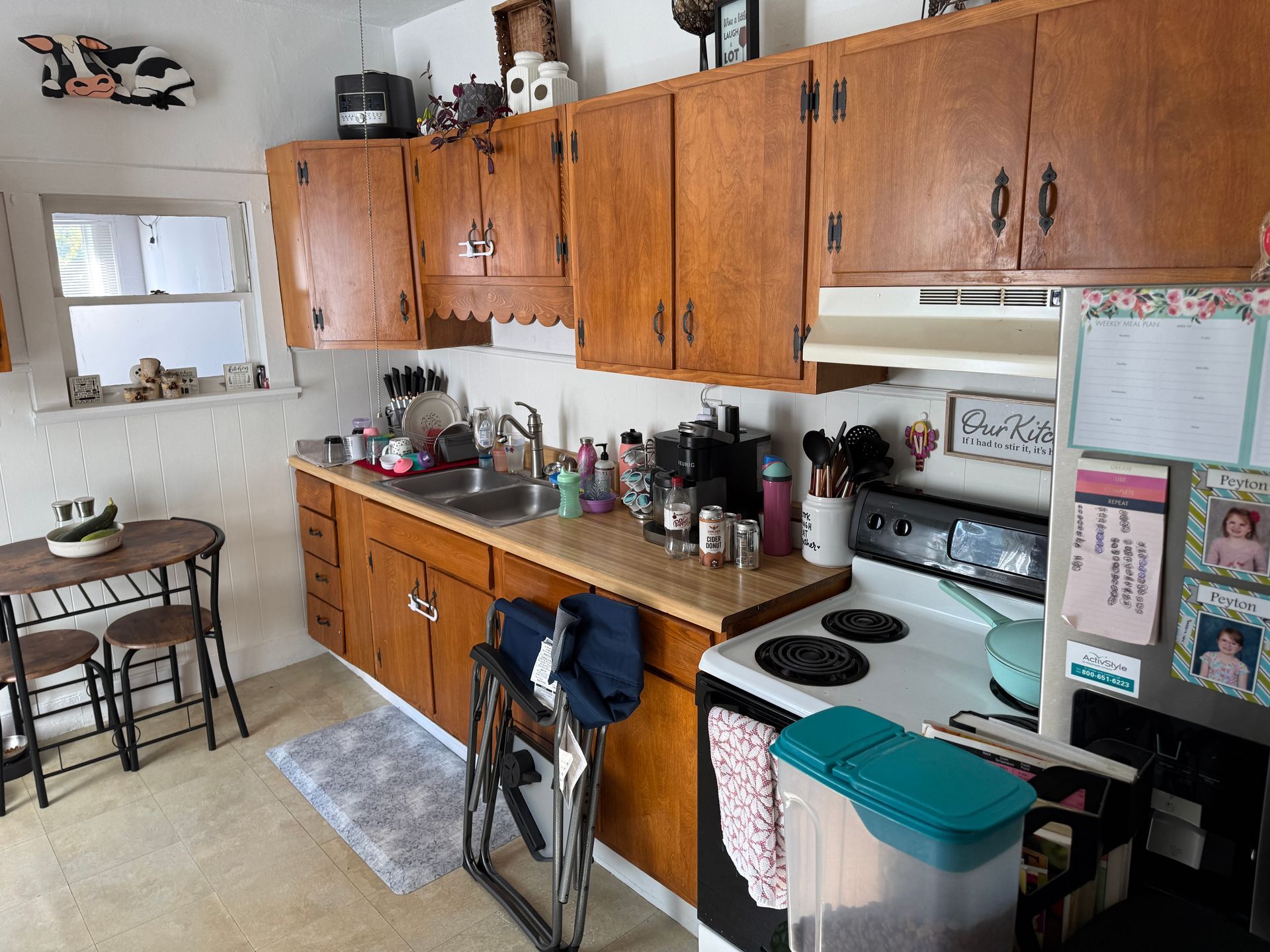 A small kitchen with wooden cabinets, appliances, and a table. A cow decoration is above the window.