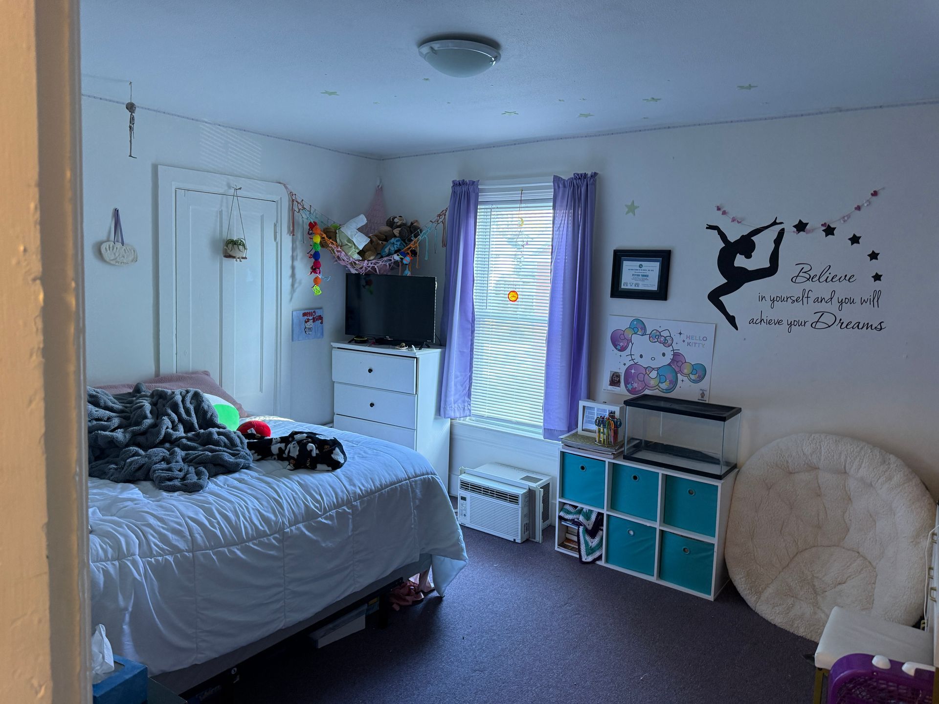 Bedroom with bed, dresser, TV, and decorative wall decals. Purple curtains and a large white rug.