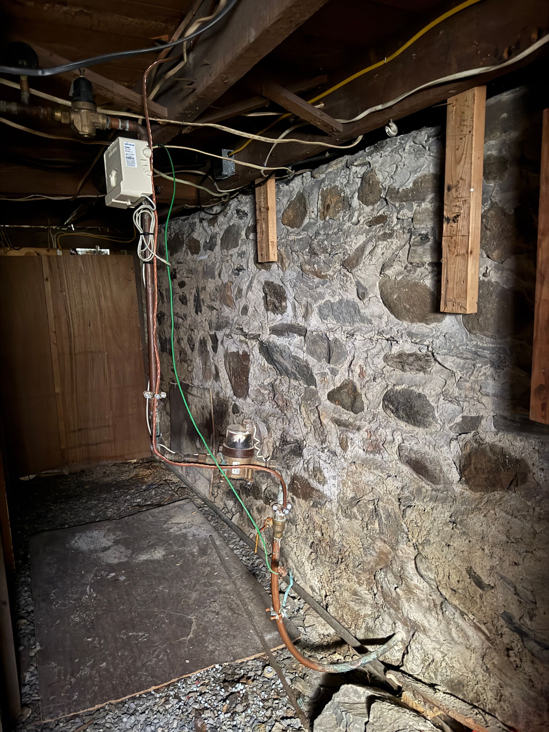 Basement with stone wall, exposed wooden beams, and electrical wiring. A metal pipe runs up the wall.