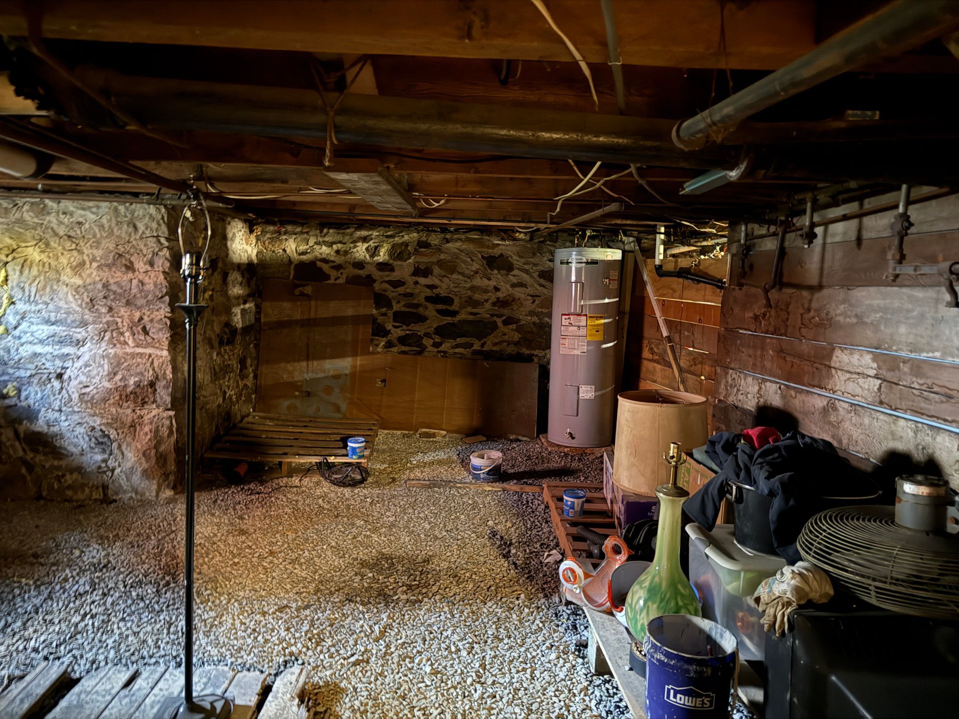 Basement interior with stone walls, gravel floor, pipes, water heater, and storage items.