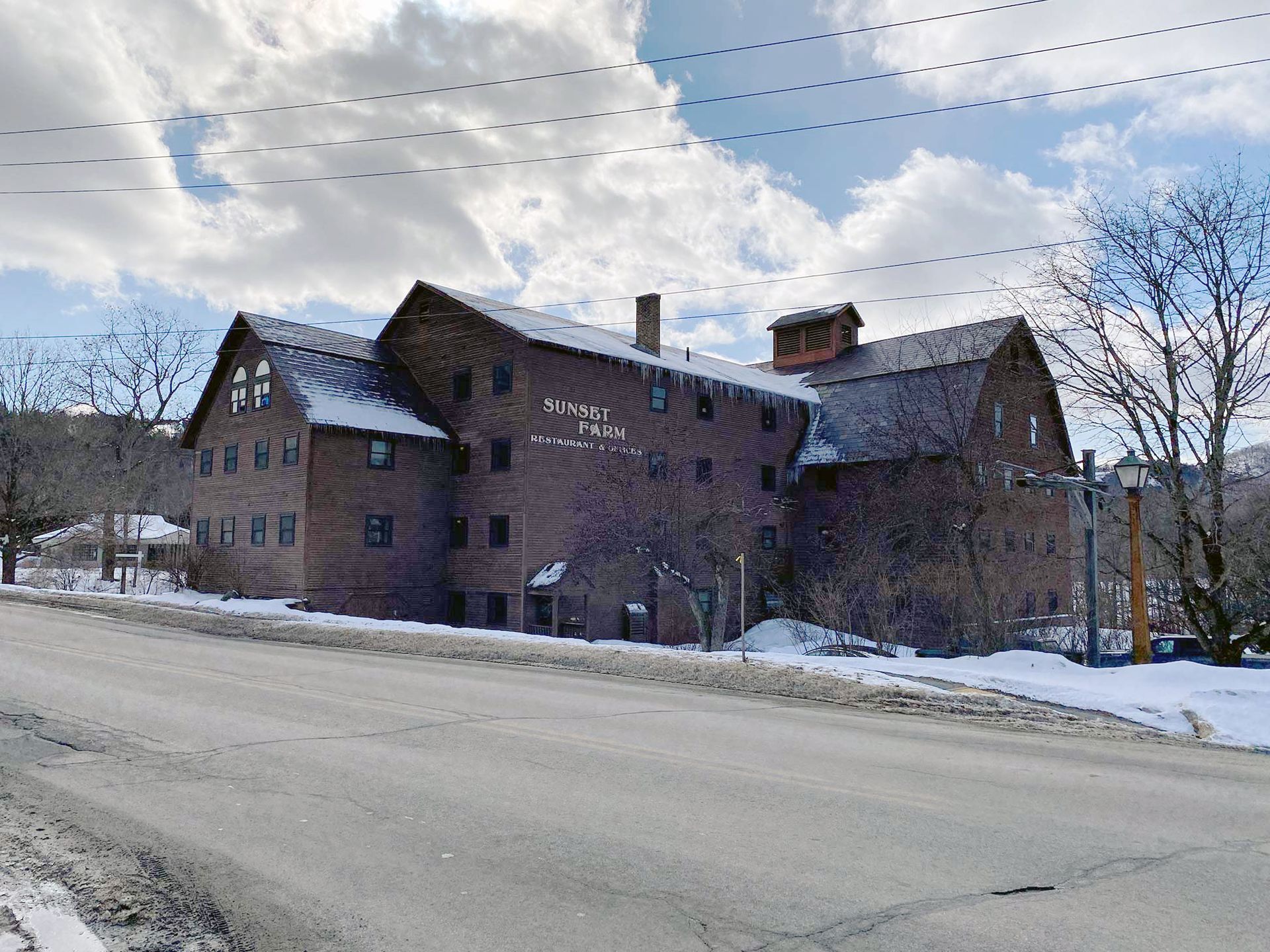Large brick building with a snow-covered roof next to a road, under a cloudy sky.