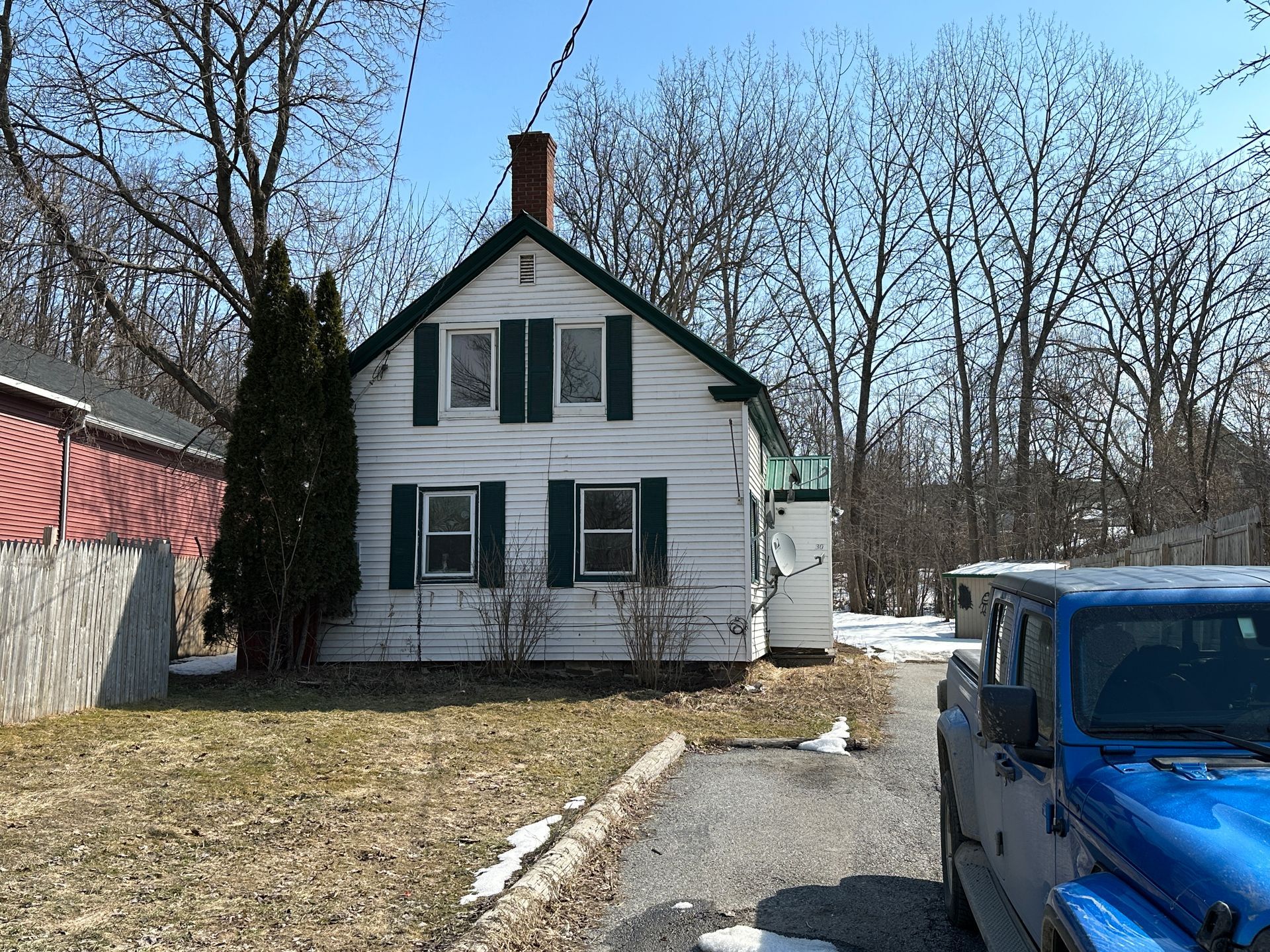 A white, two-story house with green shutters and a chimney, viewed from a driveway next to a blue Jeep on a sunny day.