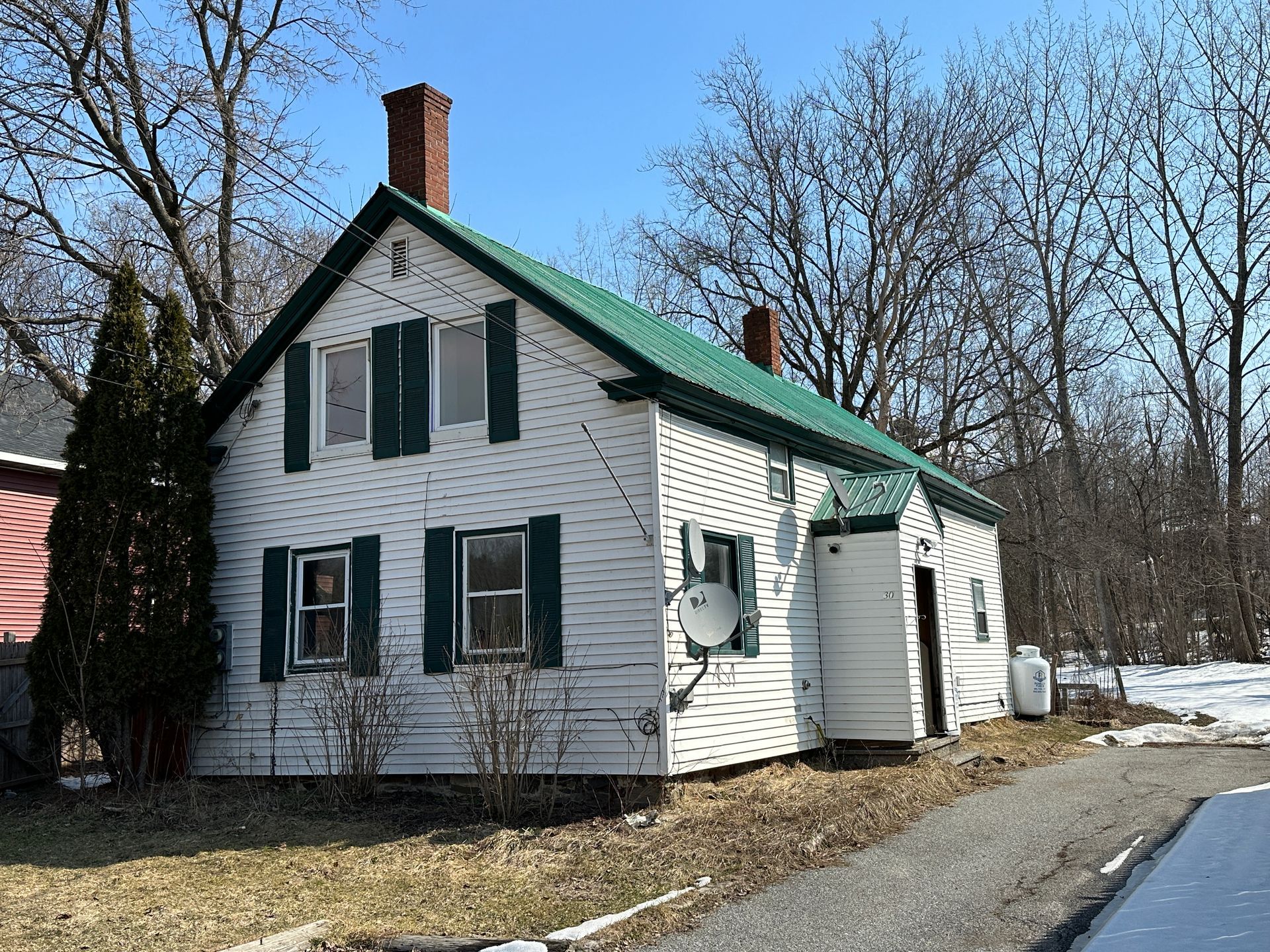 A white two-story house with a green roof, a brick chimney, and dark green shutters, set against bare trees and snow.