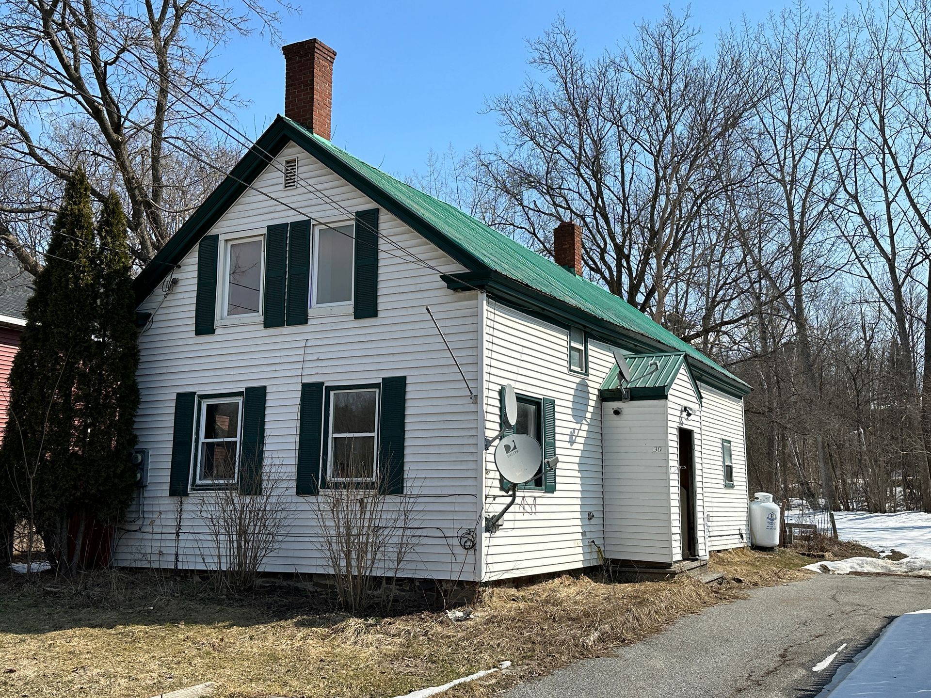 A white, two-story house with a green roof, dark shutters, and a chimney, set outdoors with trees in the background.