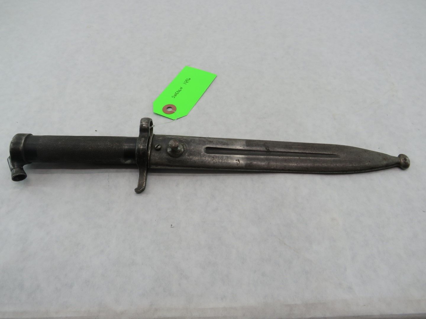 Antique, steel bayonet with dark handle and blade, lying horizontally on a light surface.