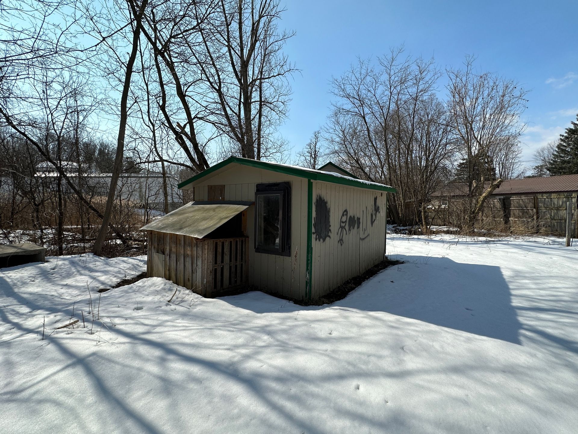 A small, weathered shed with a wooden addition stands in a snow-covered yard surrounded by bare trees under a blue sky.