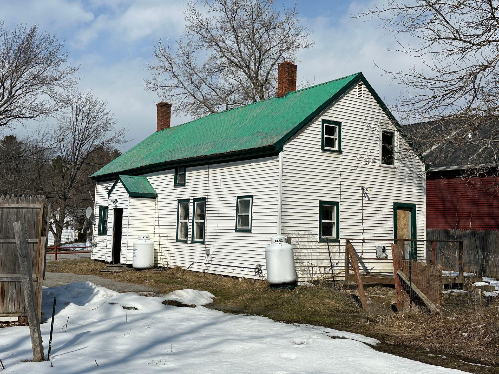 A white, one-story house with a green metal roof and two large propane tanks, set on a snowy lot with leafless trees.