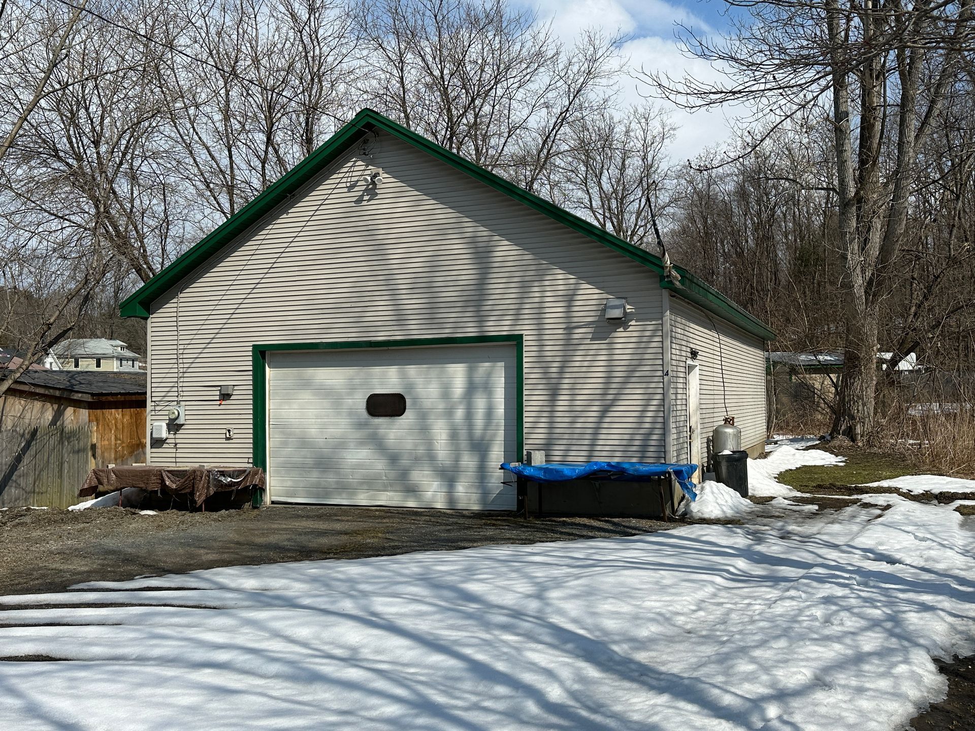 A beige garage with a green-trimmed roof, white door, and side entrance, set on a snowy, wooded lot.